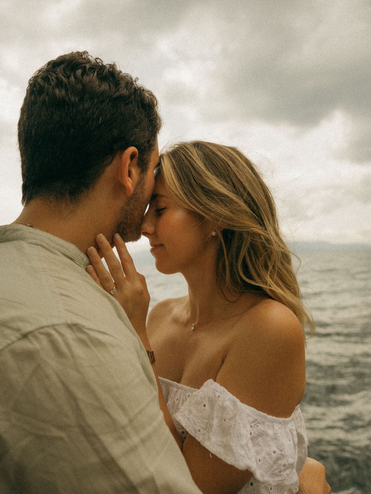 Romantic couple sharing an intimate moment by the ocean under dramatic cloudy skies, woman in white off-shoulder top.