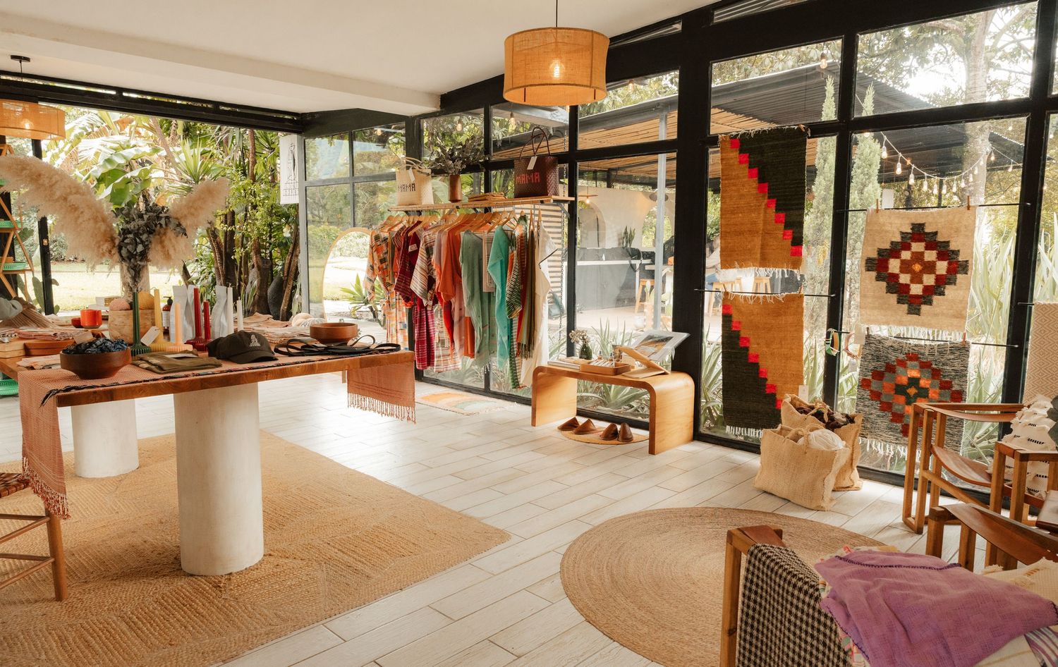 Bright boho boutique with wooden displays, colorful hanging garments, woven rugs, and rattan chairs amid lush greenery.