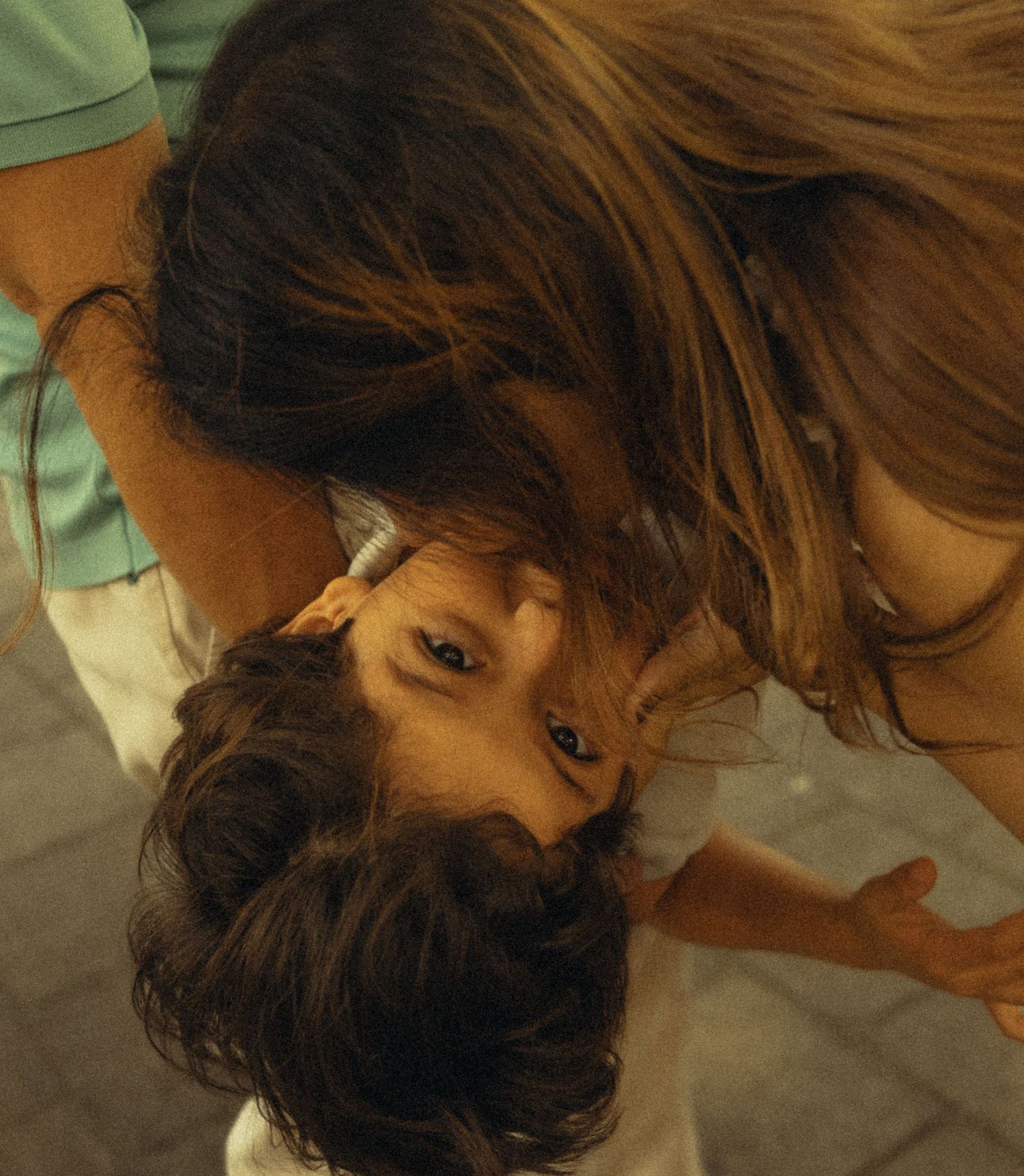 A woman with long hair bends down to kiss a young child on the top of their head outdoors.