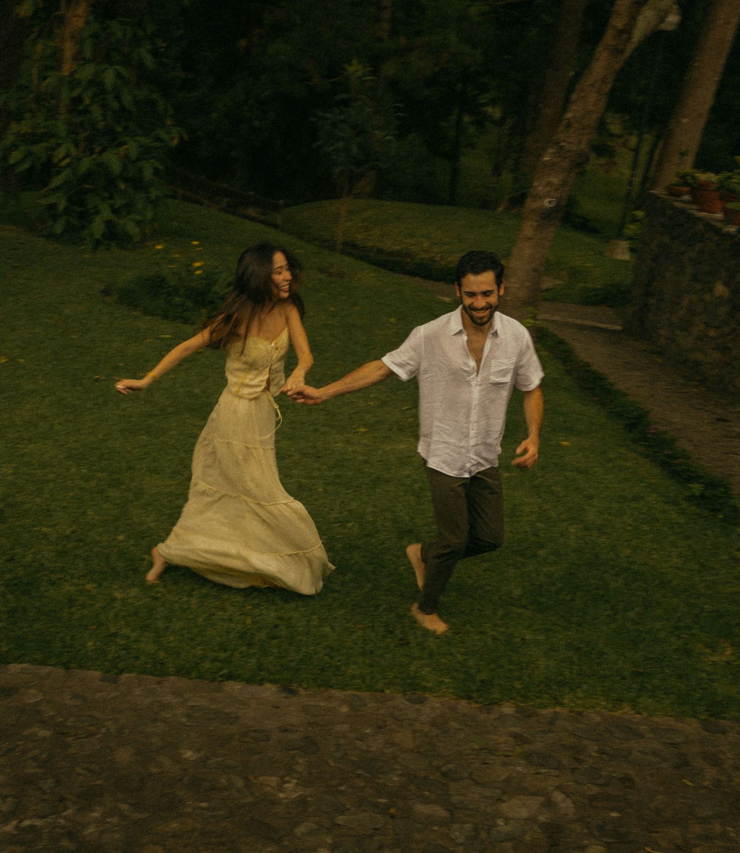 A couple runs barefoot through lush green grass, holding hands, woman in flowing cream dress, man in white shirt.