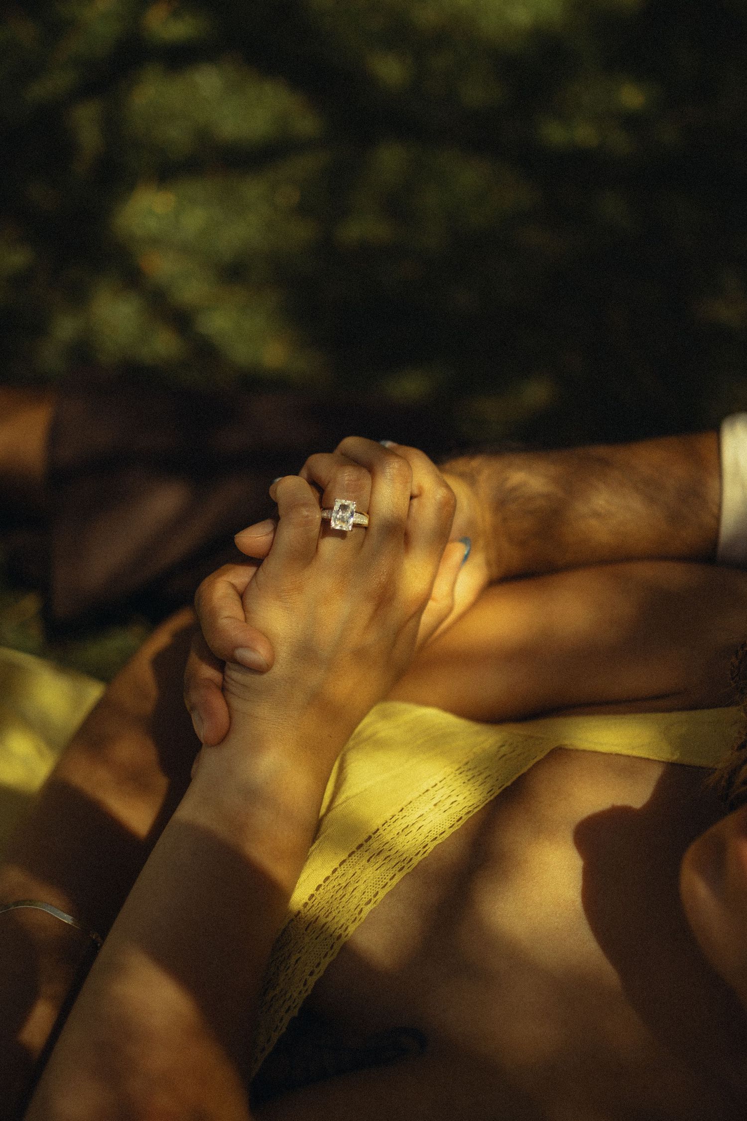 Two hands clasped together with a diamond engagement ring in warm sunlight outdoors.