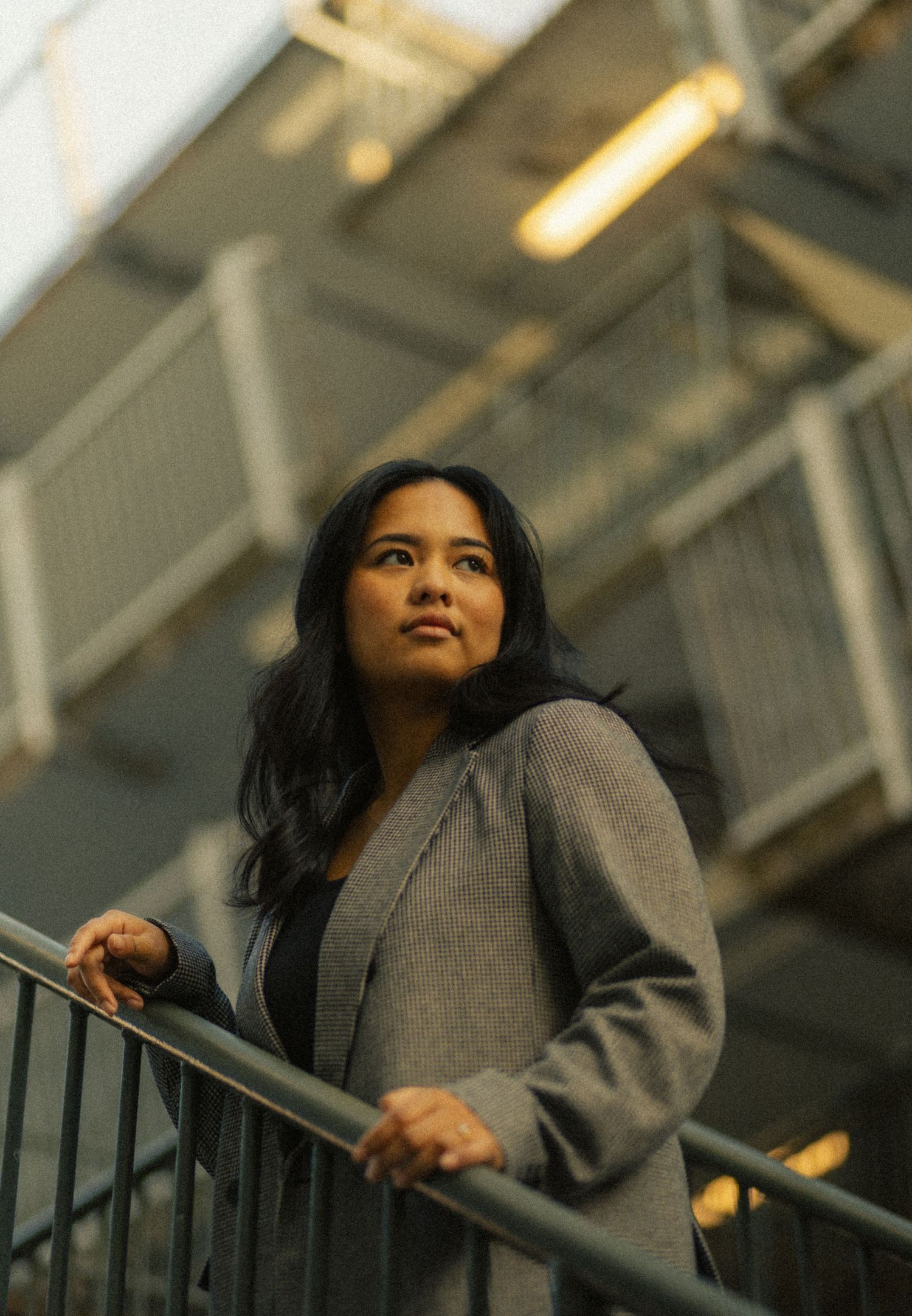 A young woman in a gray blazer stands on metal stairs, gazing upward in an urban industrial setting.