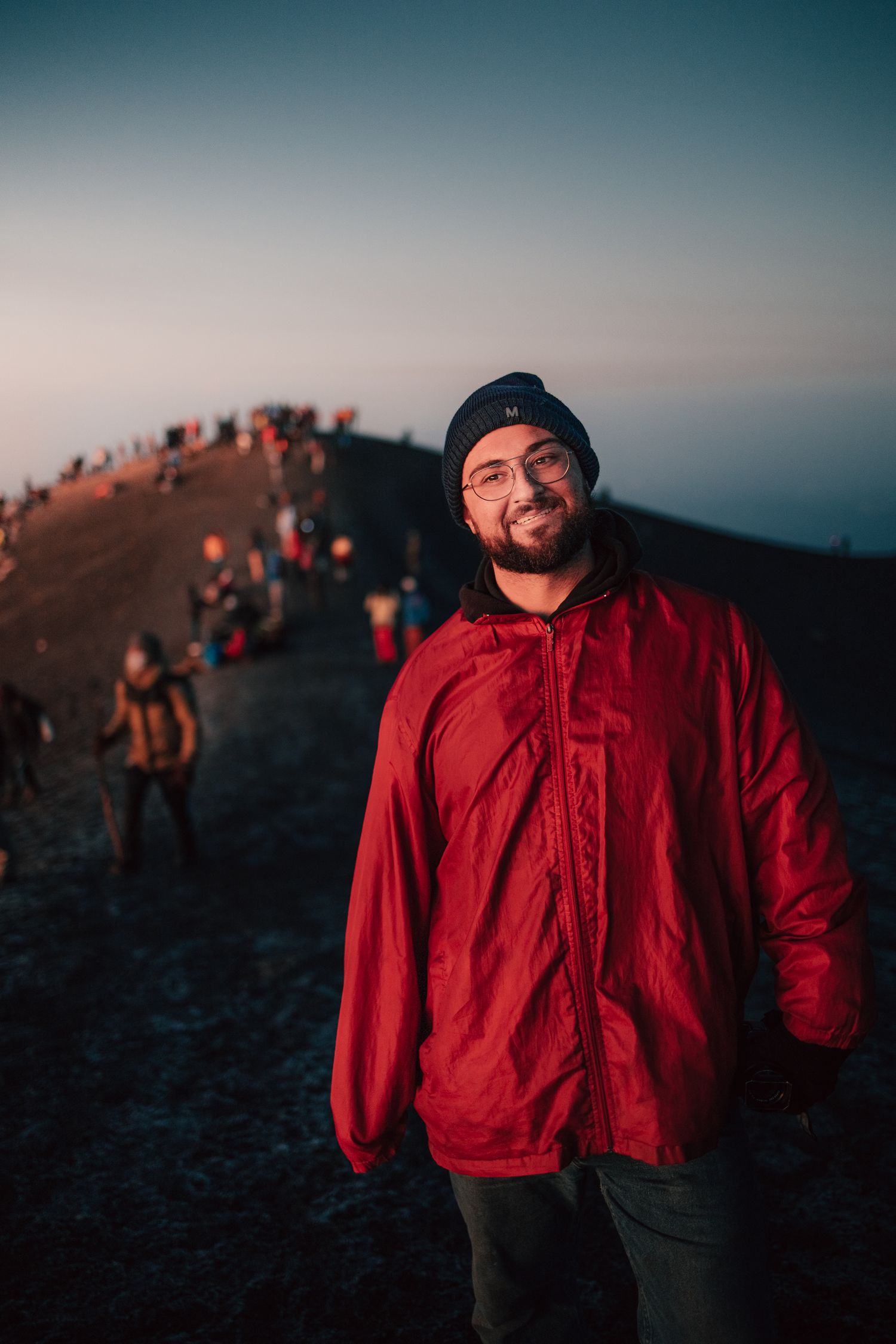 Smiling man in red jacket and beanie hat standing on mountain summit at dusk with hikers in background.