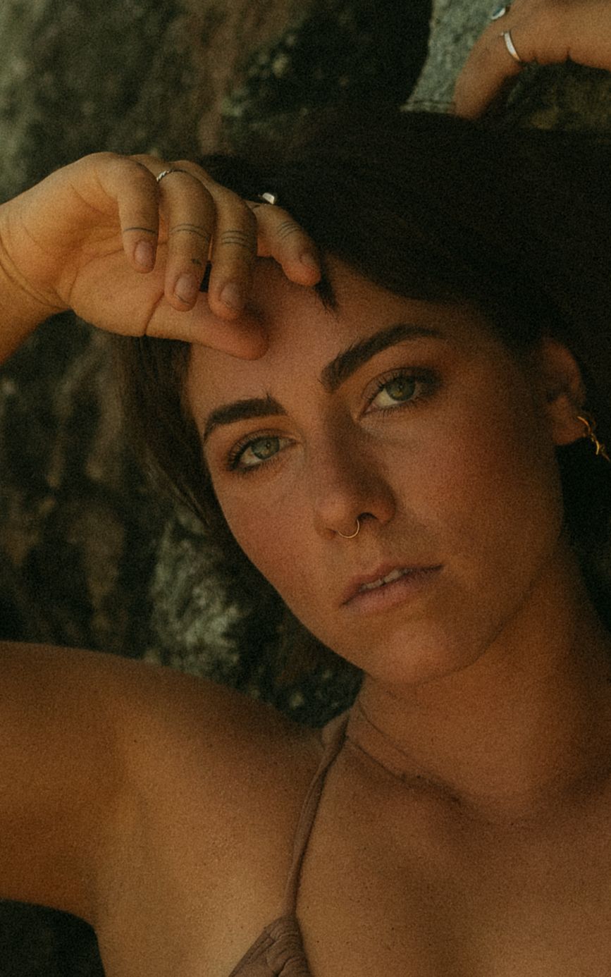 Young woman with green eyes, nose ring, and rings on fingers poses outdoors against rocky background in warm light.