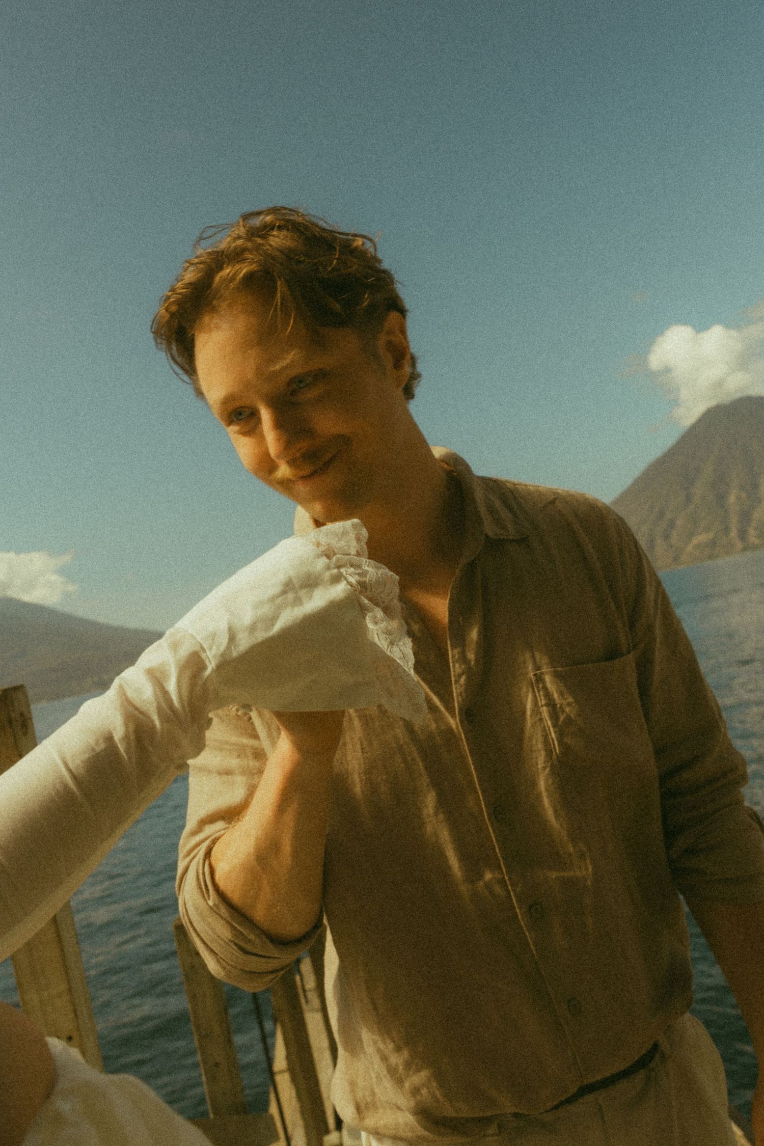 Smiling young man in linen shirt leaning on wooden railing with scenic lake and mountains behind him.
