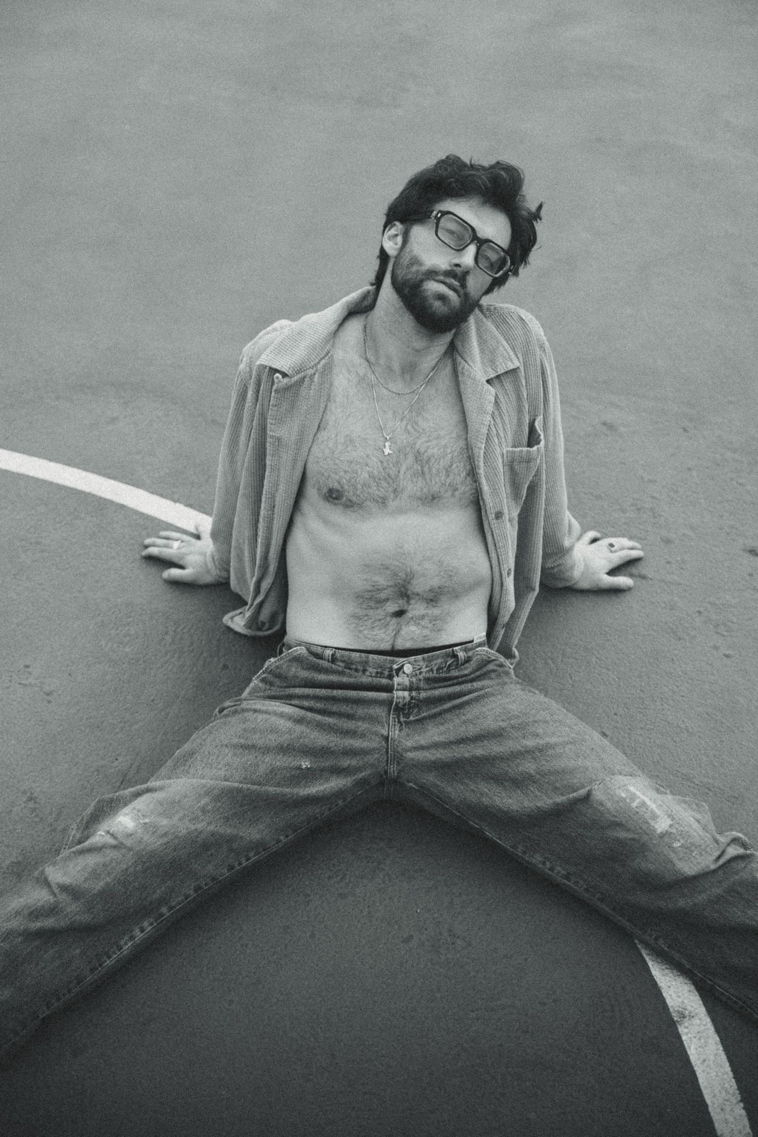 Black and white photo of bearded man in open shirt and jeans lying on pavement wearing glasses.
