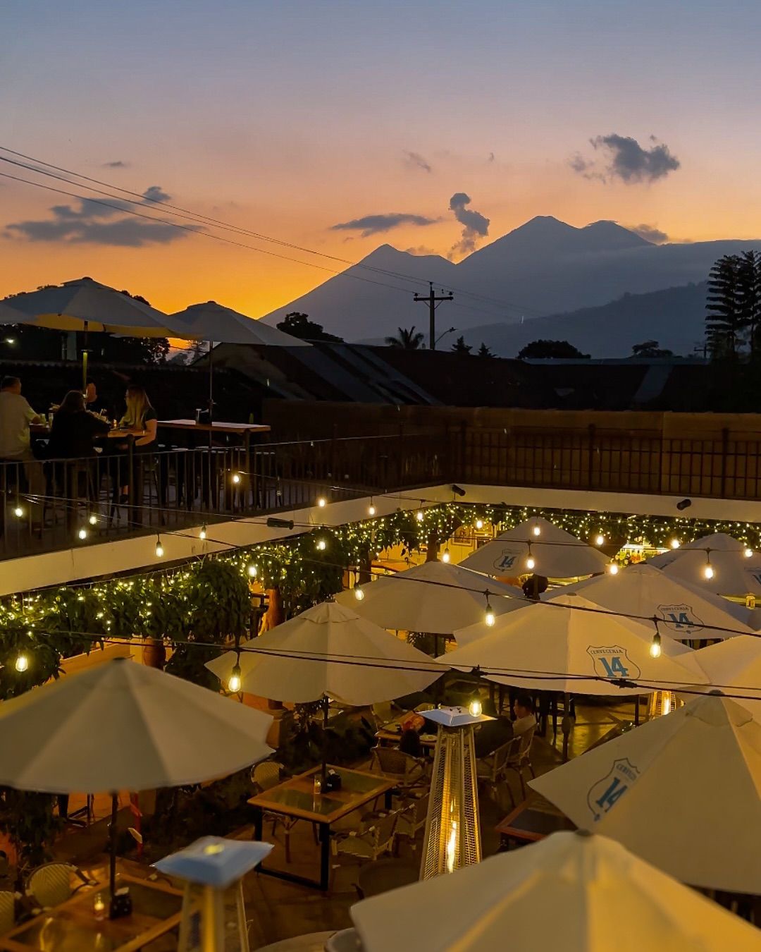 Outdoor rooftop restaurant with string lights and umbrellas at sunset, volcanic mountain silhouettes in the background.