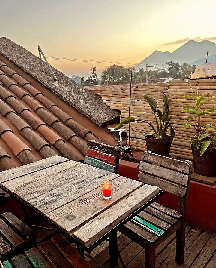 Rooftop terrace with wooden table and chairs, terracotta tiles, potted plants, and volcanic mountain sunset view.