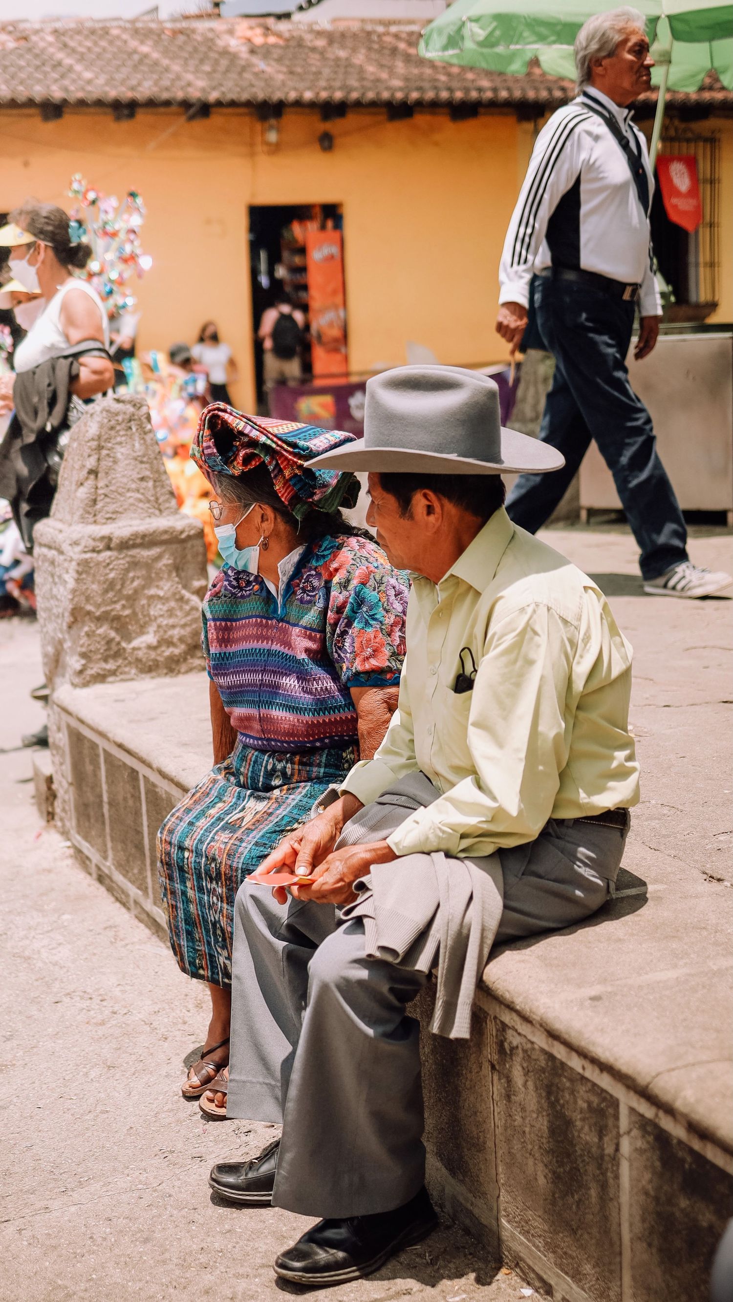 Elderly Guatemalan couple in traditional clothing sitting on a stone ledge at a busy outdoor market.