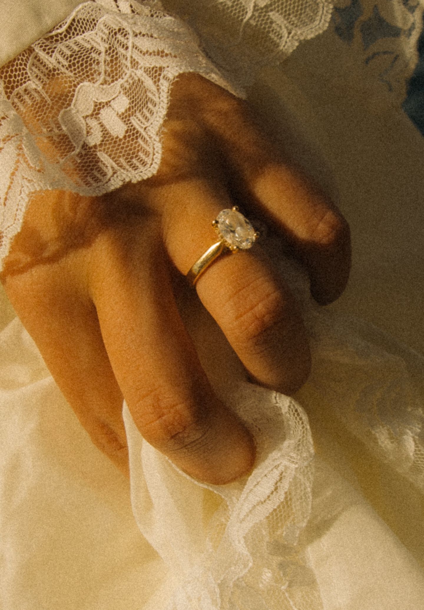 Close-up of a hand wearing a gold solitaire diamond engagement ring resting on white lace fabric.