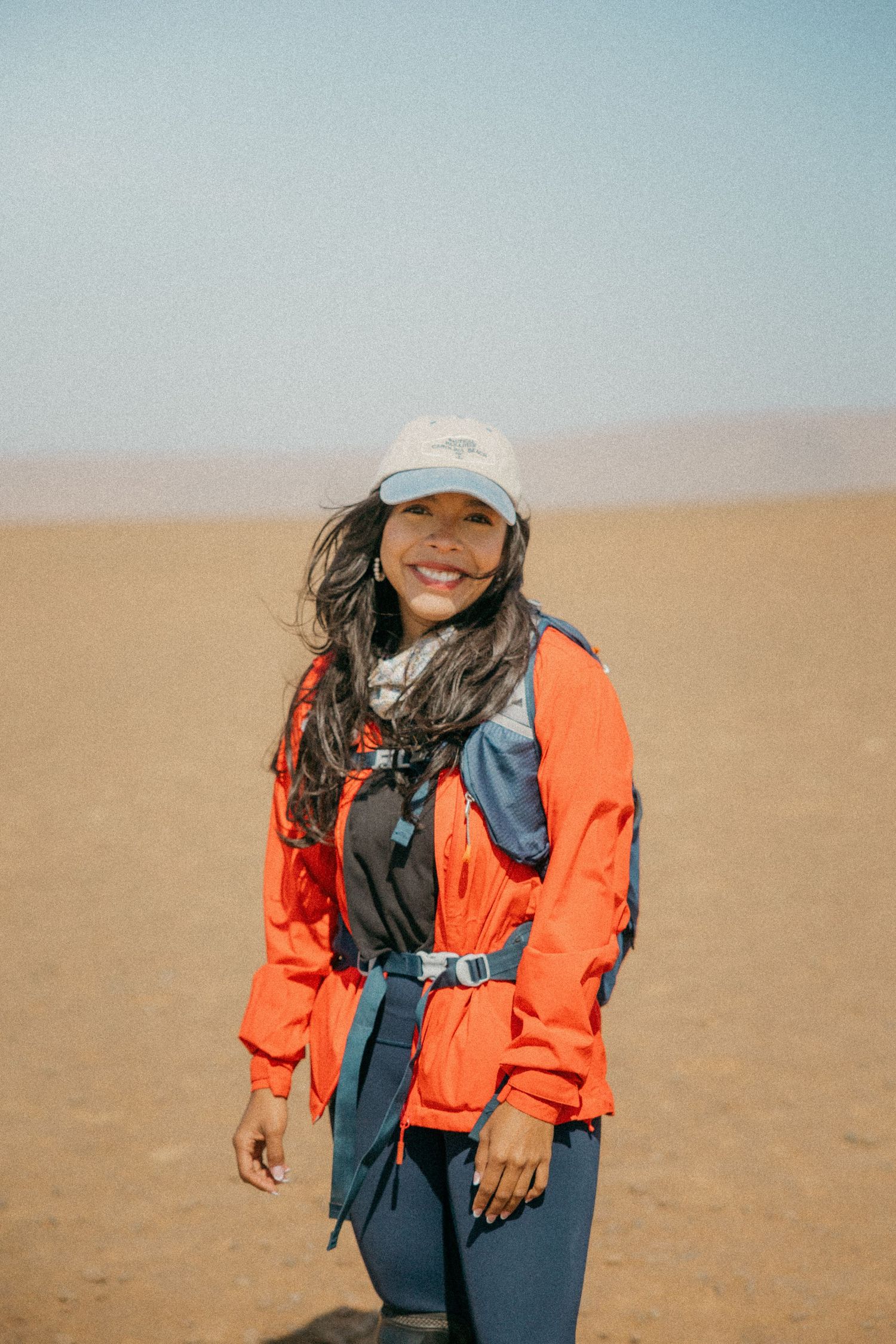 Smiling woman in orange jacket and white cap hiking in a vast desert landscape on a sunny day.