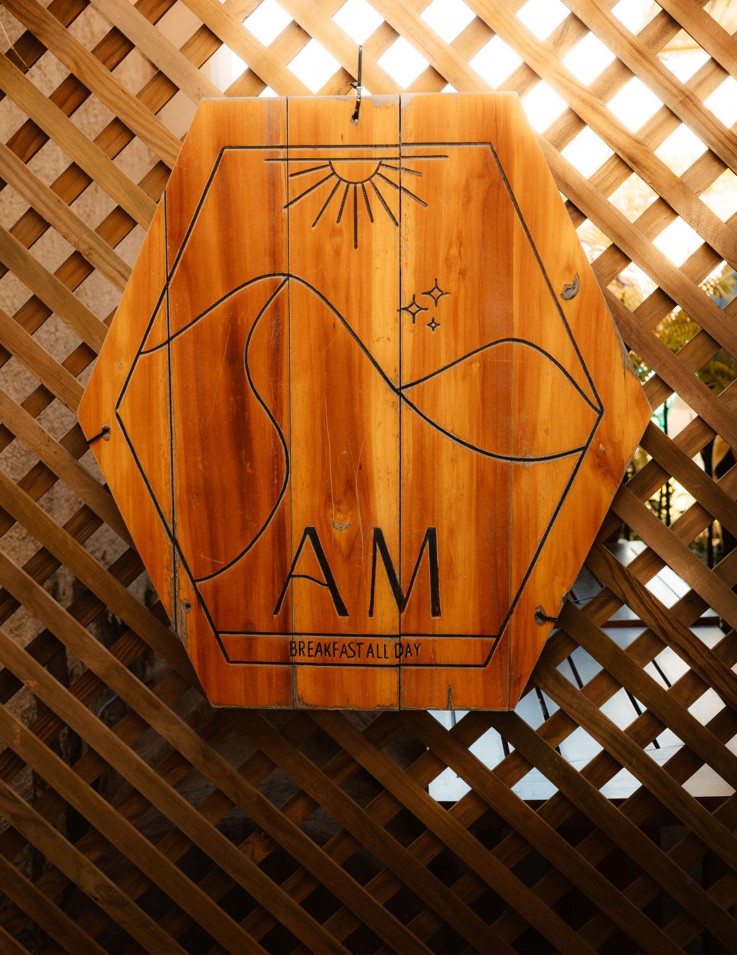 Hexagonal wooden sign reading AM Breakfast All Day, hanging on a lattice wall.