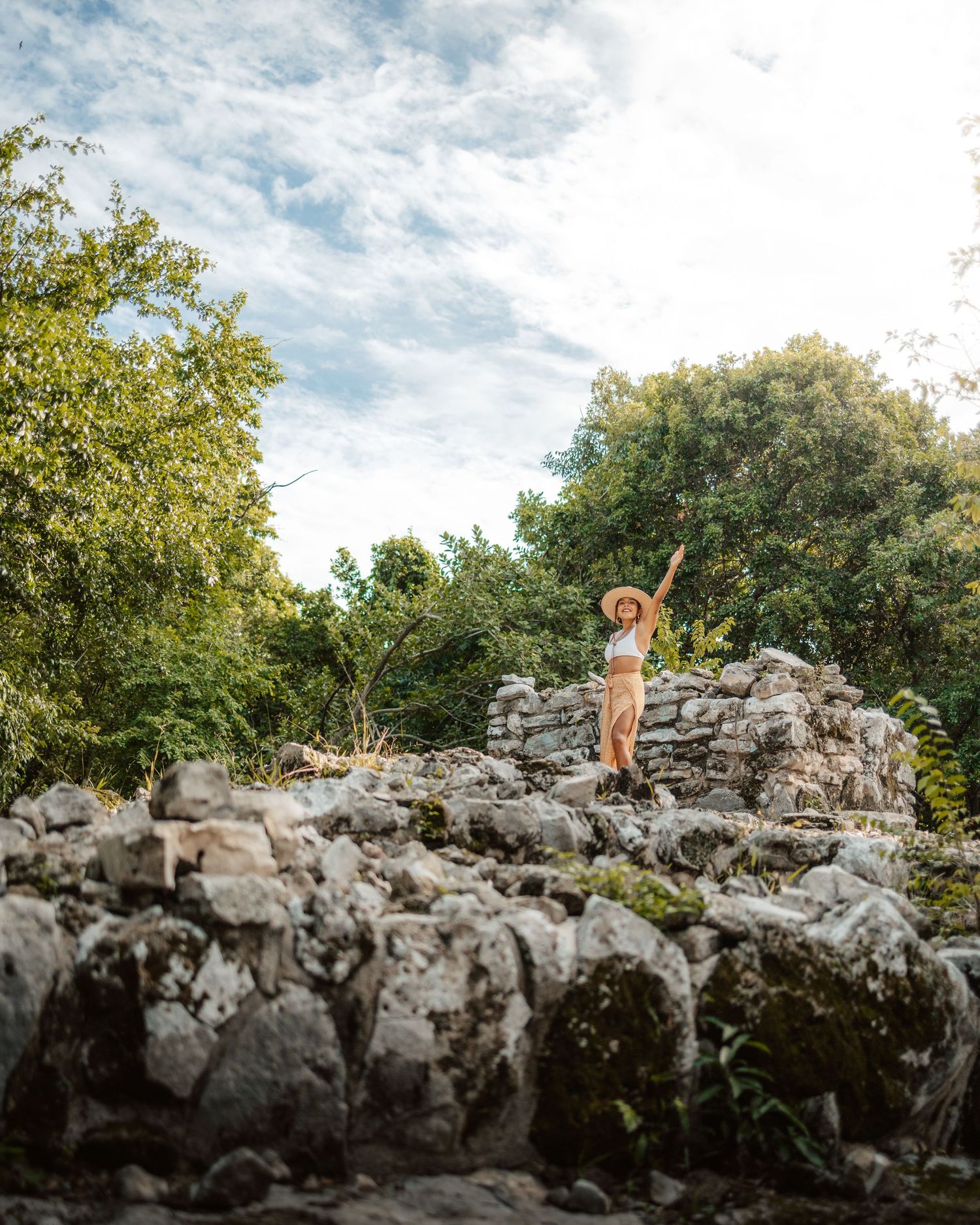 Woman in hat and crop top stands atop ancient stone ruins surrounded by lush green trees under a cloudy sky.