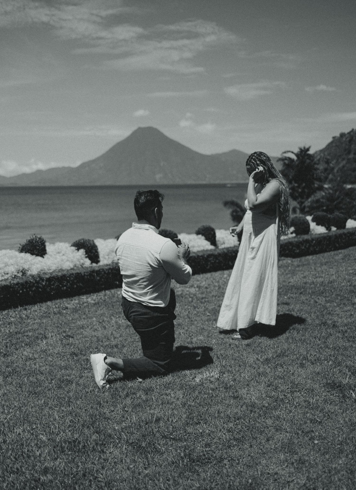 Man proposes on one knee to woman in white dress by a lake with volcanic mountain backdrop in black and white.
