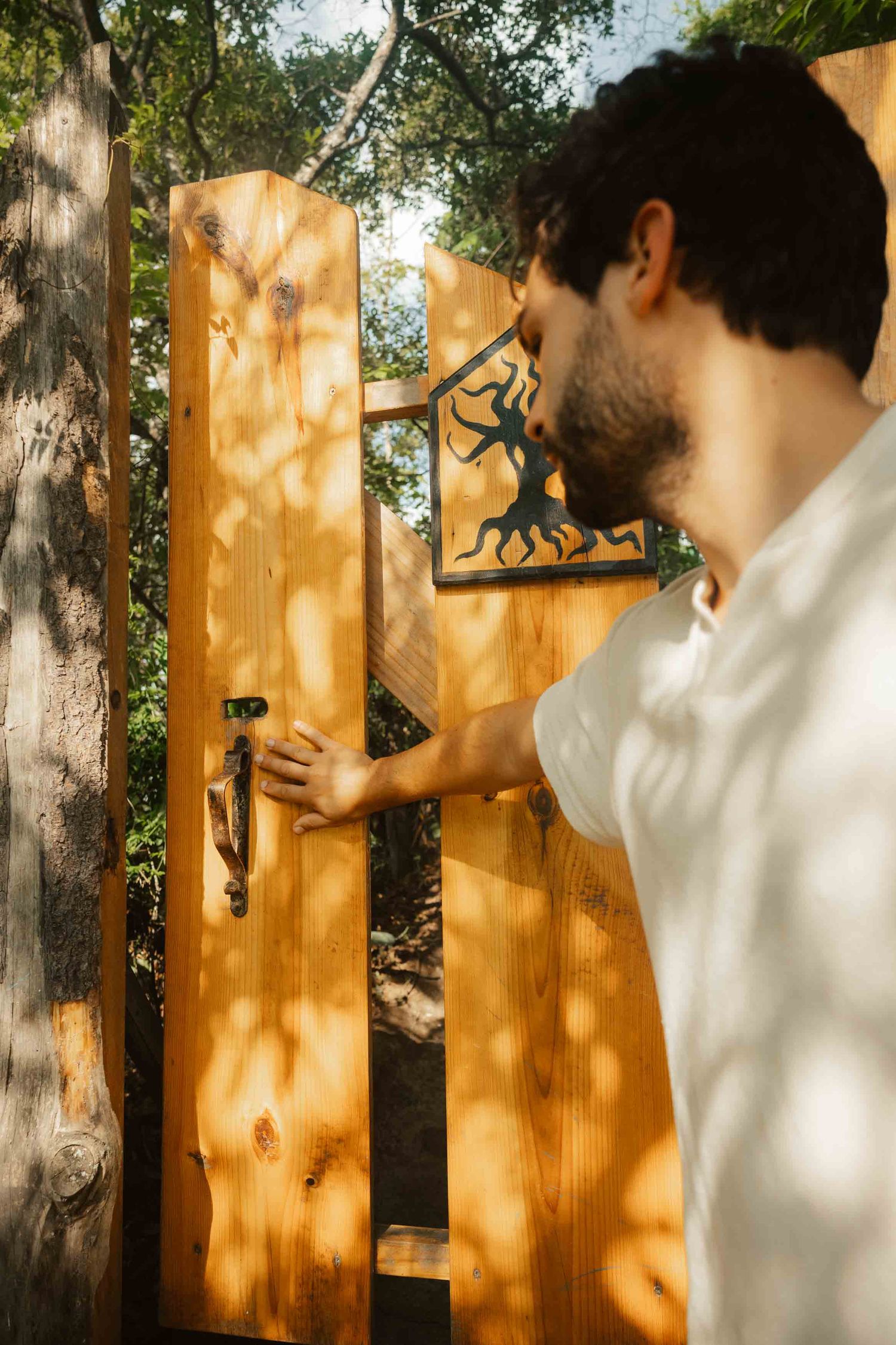 A bearded man in white shirt opening a wooden garden gate with decorative metal art panel outdoors.