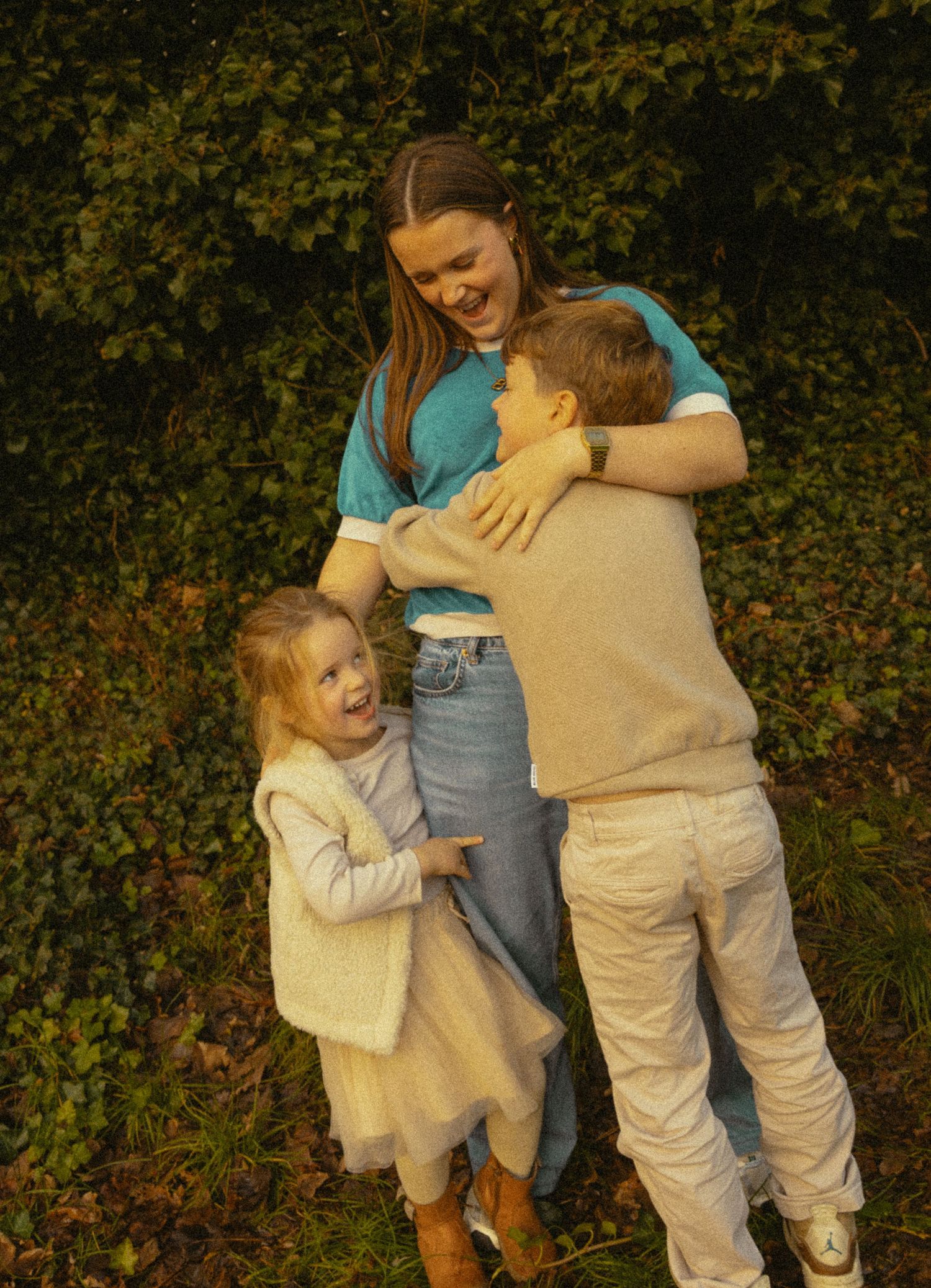 A young woman laughs while hugging two small children outdoors near a lush green hedge.