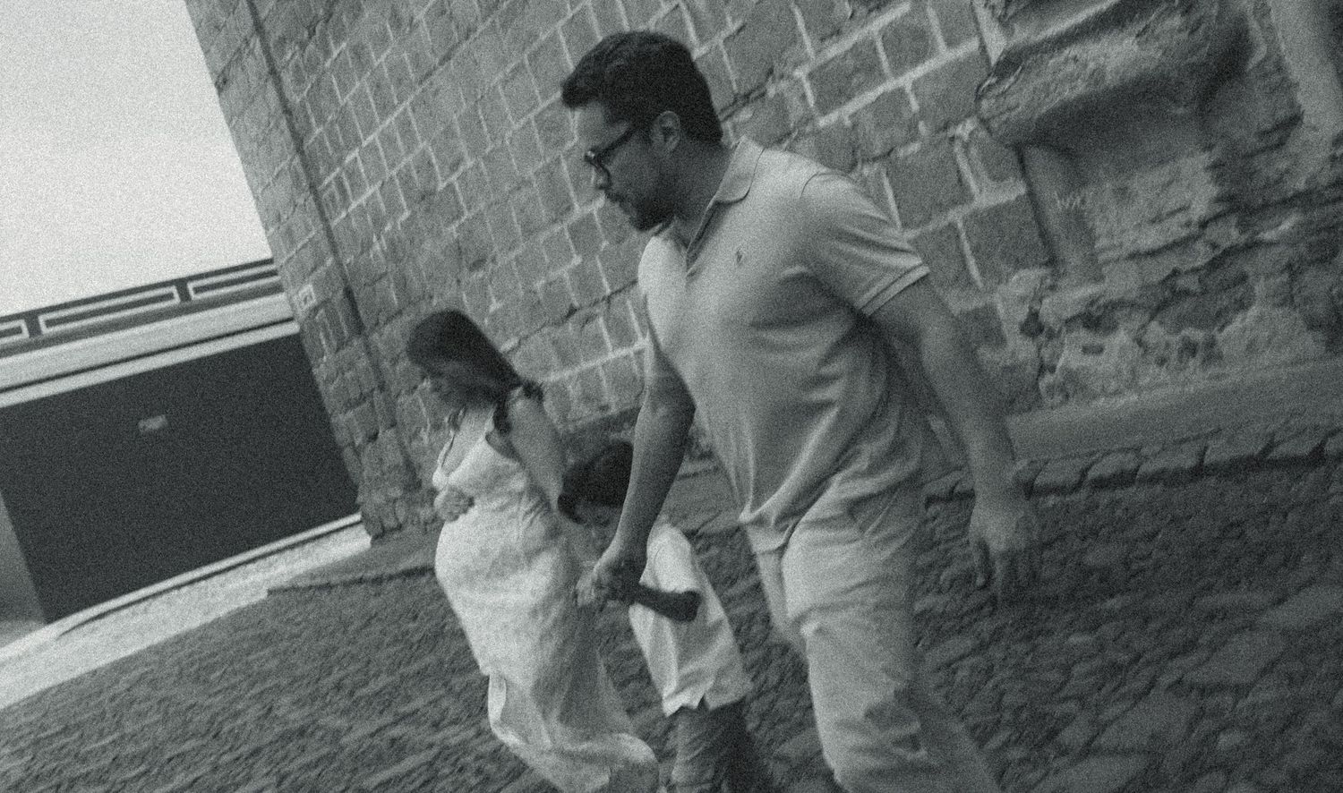 Black and white photo of a man holding a child's hand walking along a cobblestone path beside a brick wall.
