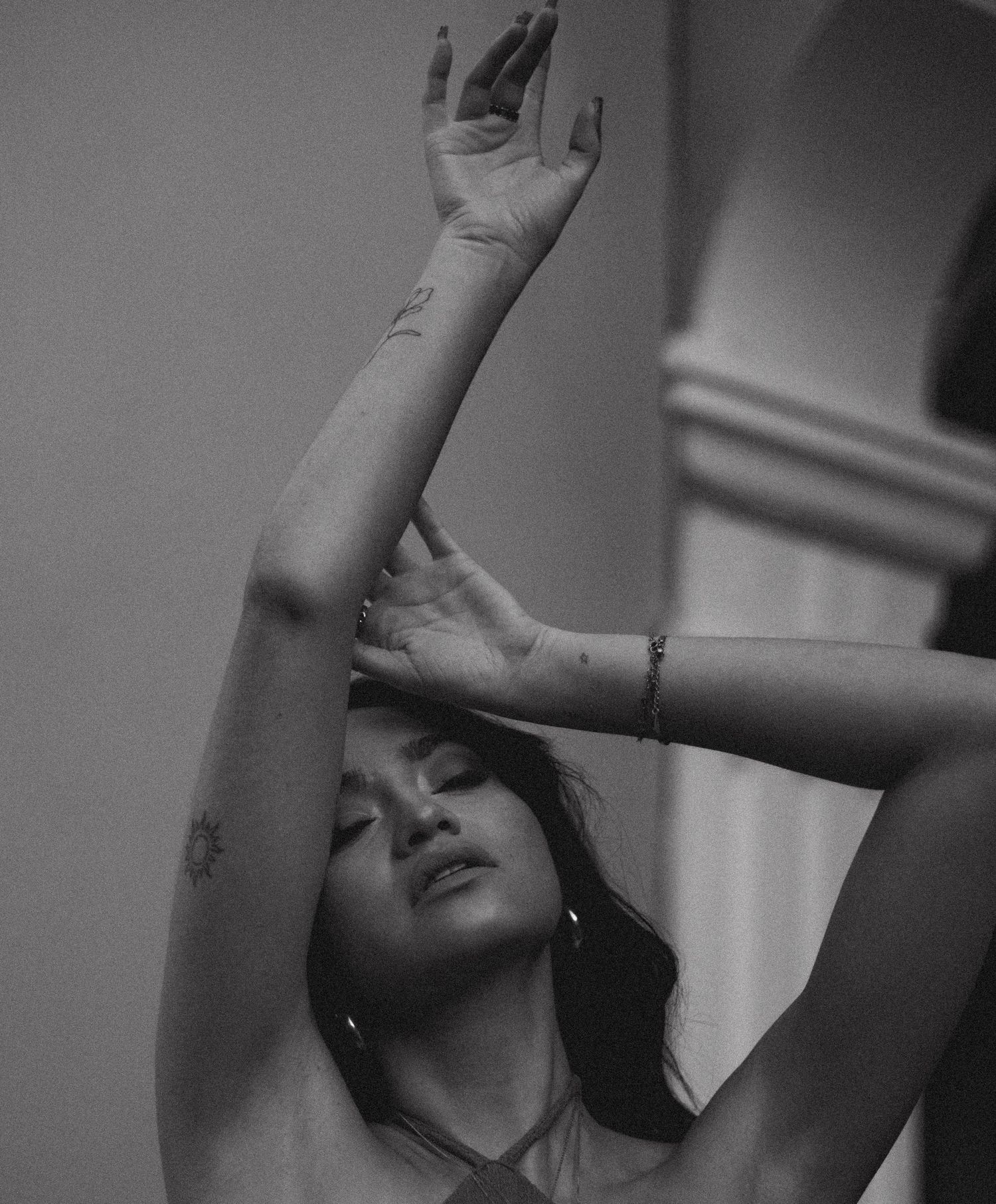 Black and white photo of a woman with arms raised, eyes closed, showing tattoos and a bracelet.
