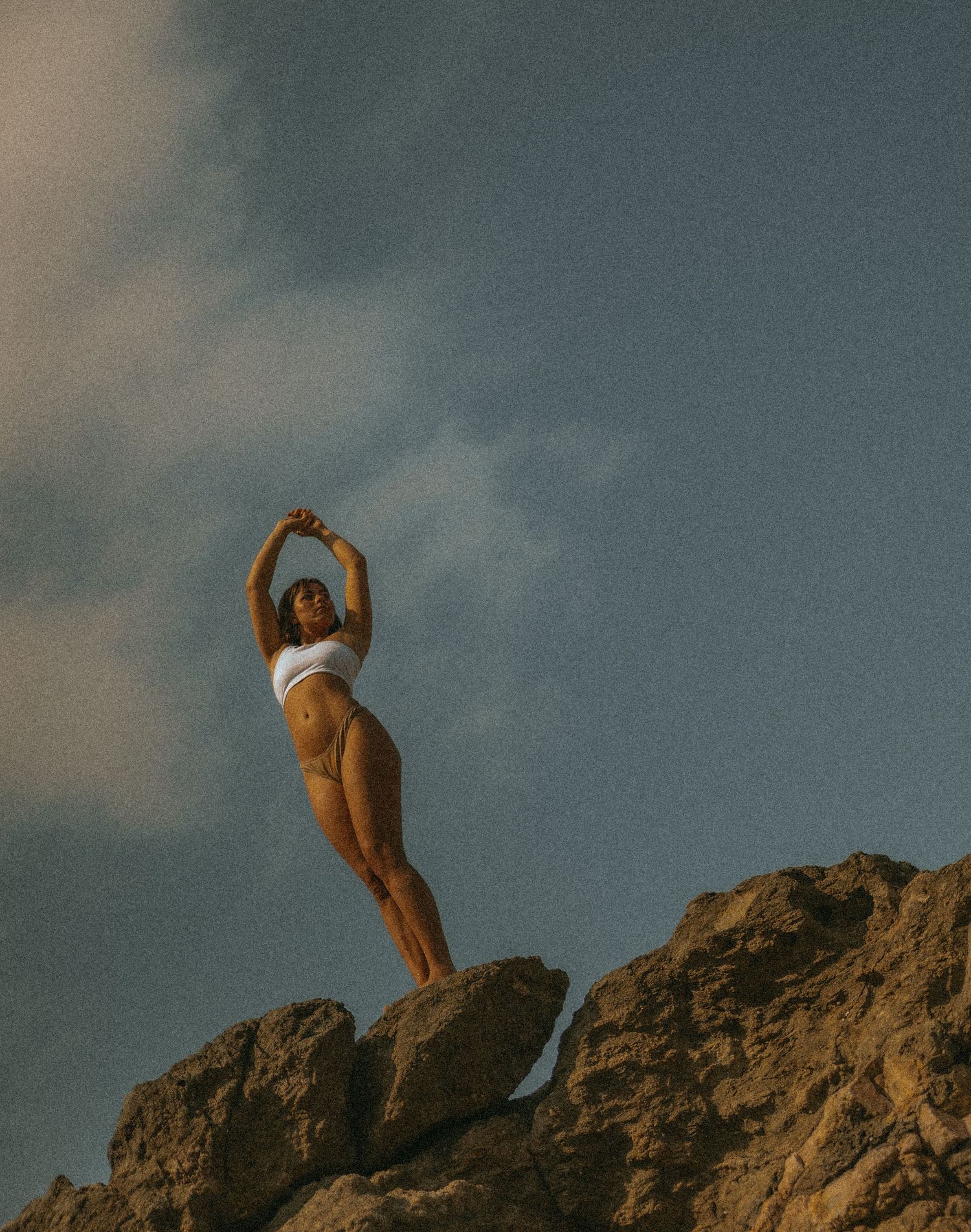Woman in white swimsuit posing triumphantly atop rocky cliff against dramatic blue sky.