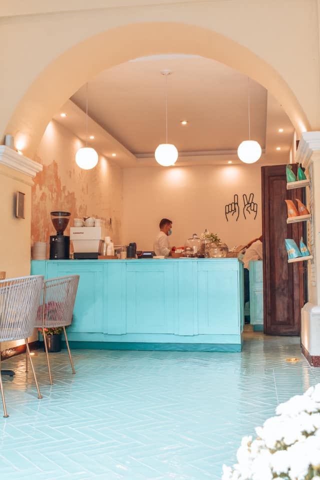 Bright cafe interior with turquoise counter, tiled floor, arch doorway, pendant lights, and barista working behind the bar.