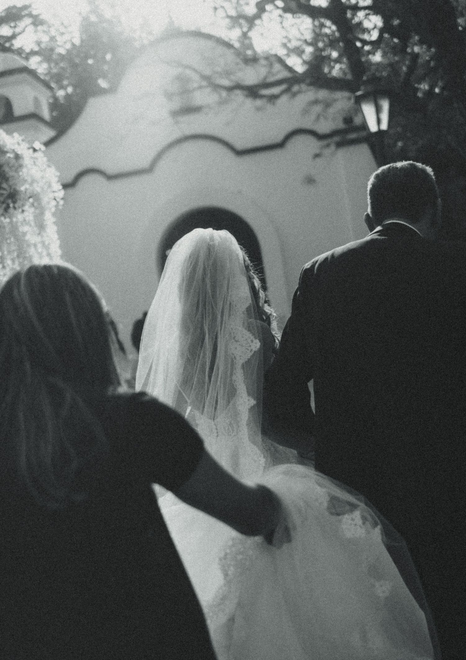 Black and white photo of a bride in a veil and groom walking toward a white chapel entrance on their wedding day.