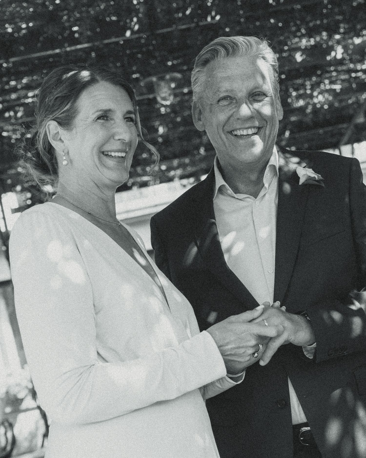Black and white photo of a smiling couple holding hands outdoors, dressed in formal attire at a celebration.