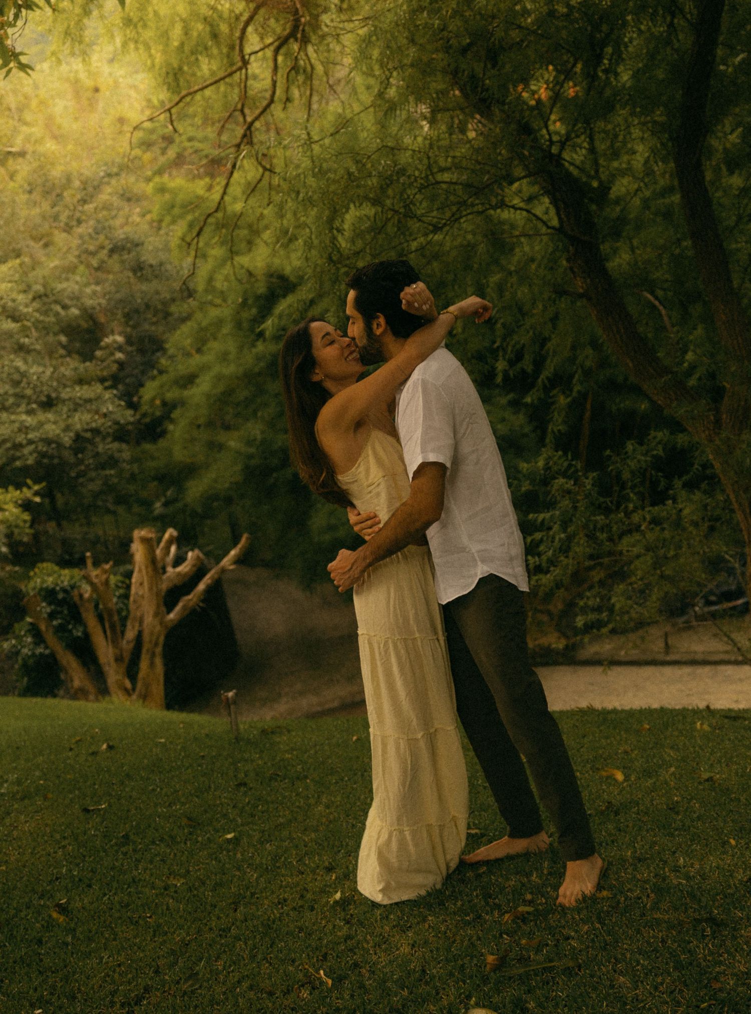 A couple embraces and dances romantically in a lush green garden surrounded by trees.
