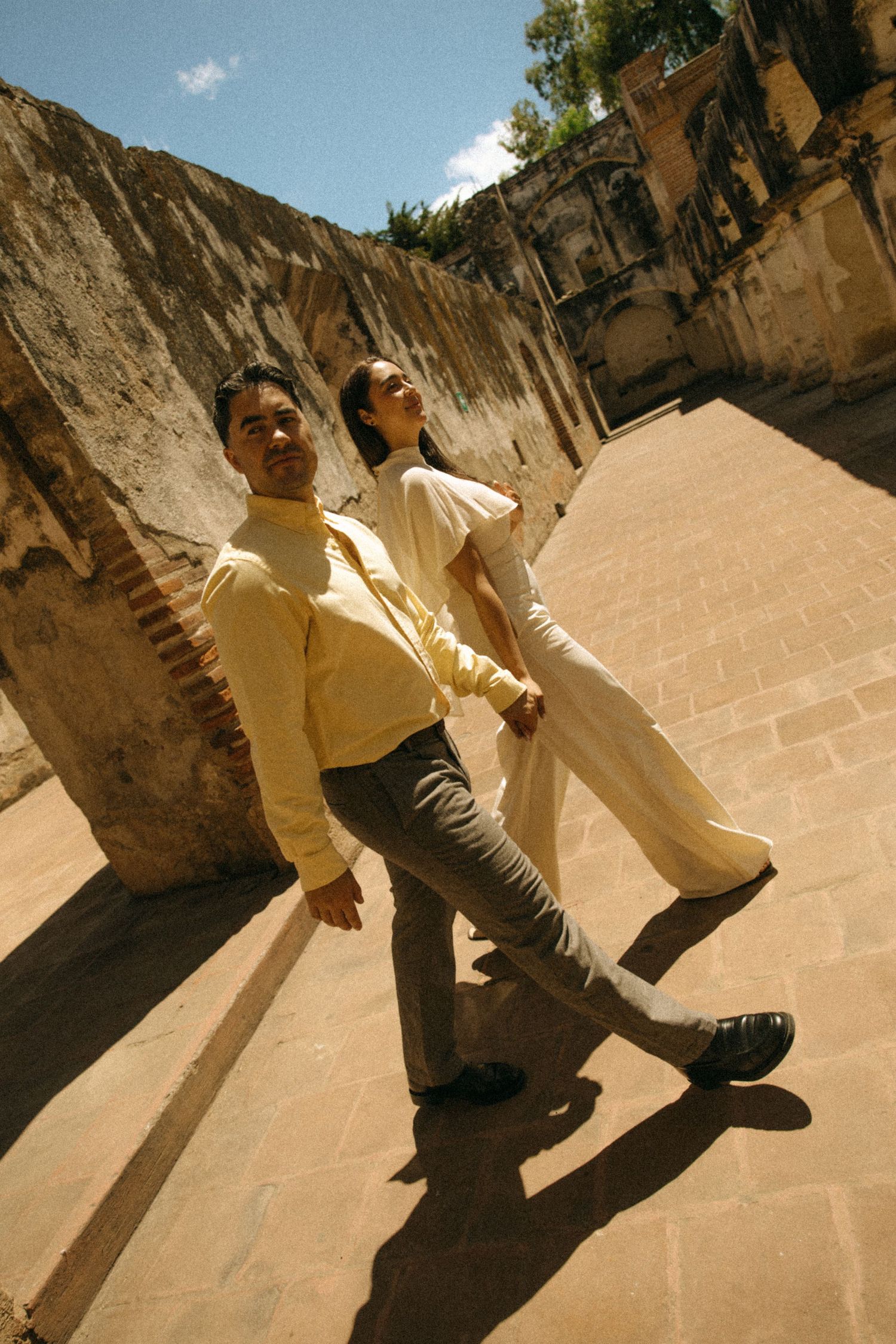 A couple in matching cream outfits stands hand in hand on a brick-paved path beside weathered stone ruins under a blue sky.