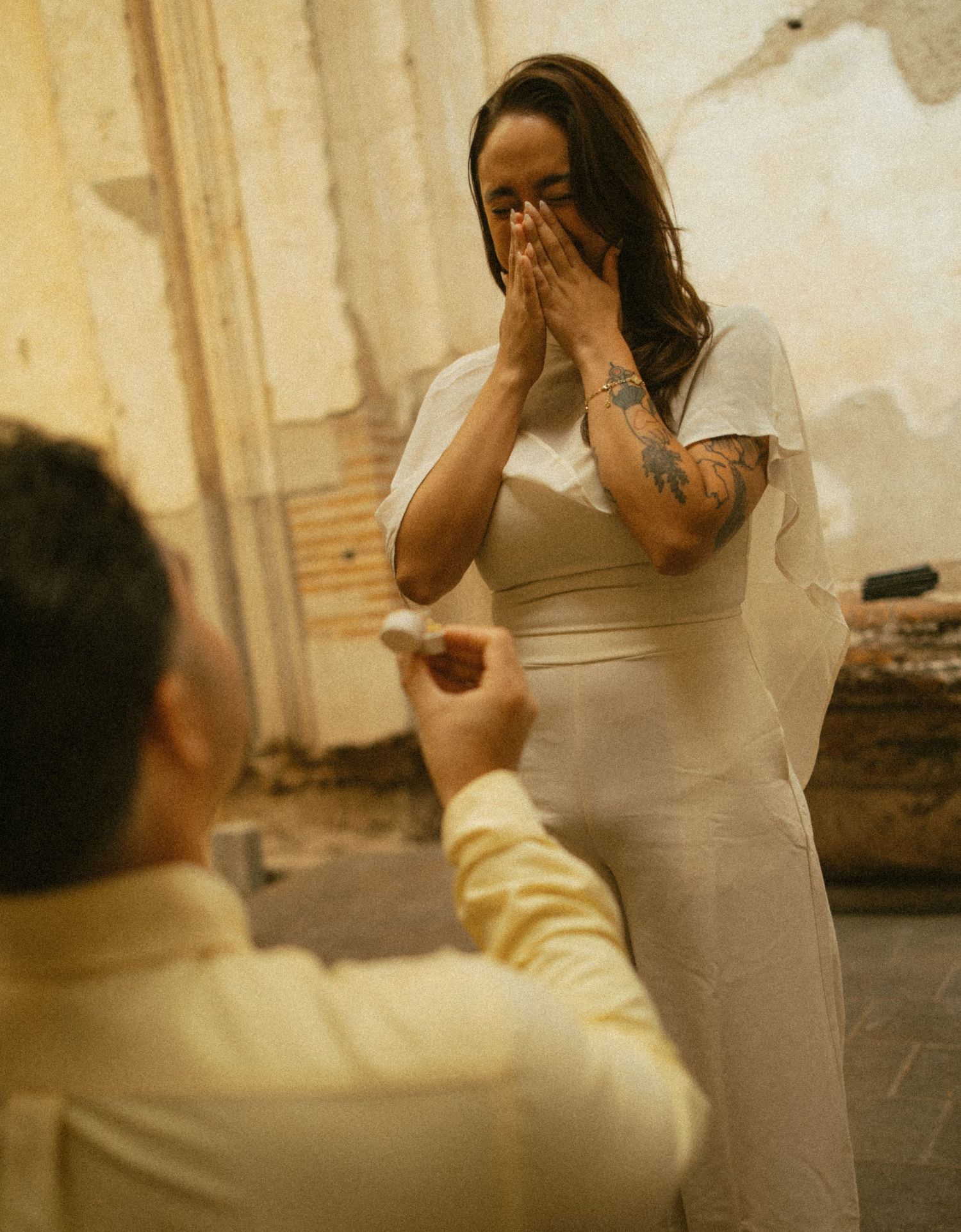 Woman in a white dress covers her face with tearful joy as a man in a cream shirt kneels and offers a ring in a rustic setting.
