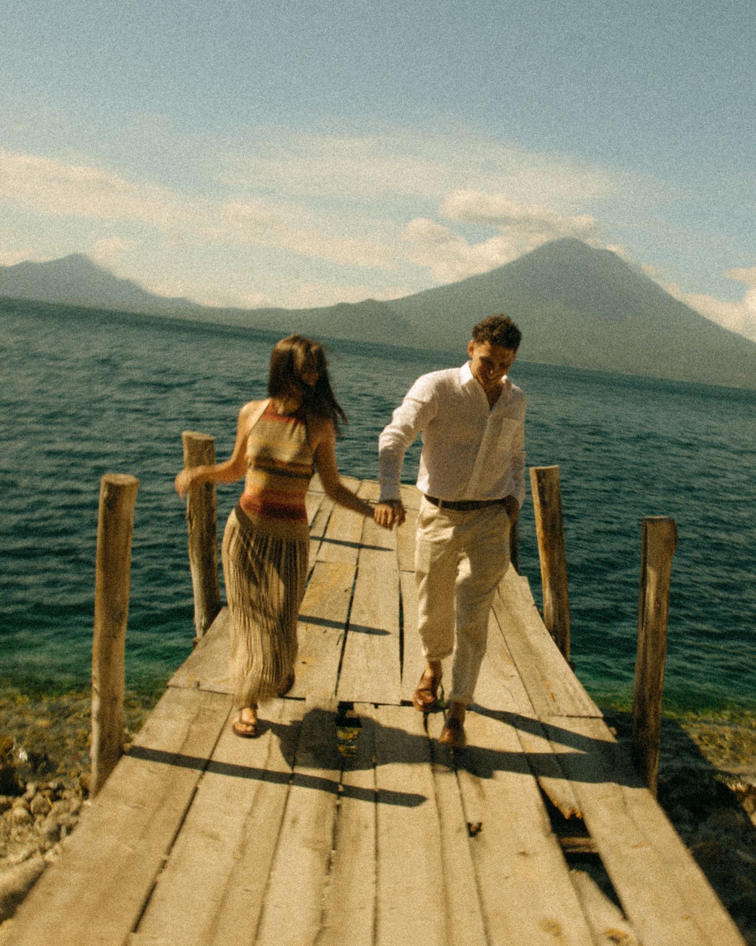Couple holding hands walking on a wooden dock with a volcanic lake and mountain backdrop under a blue sky.