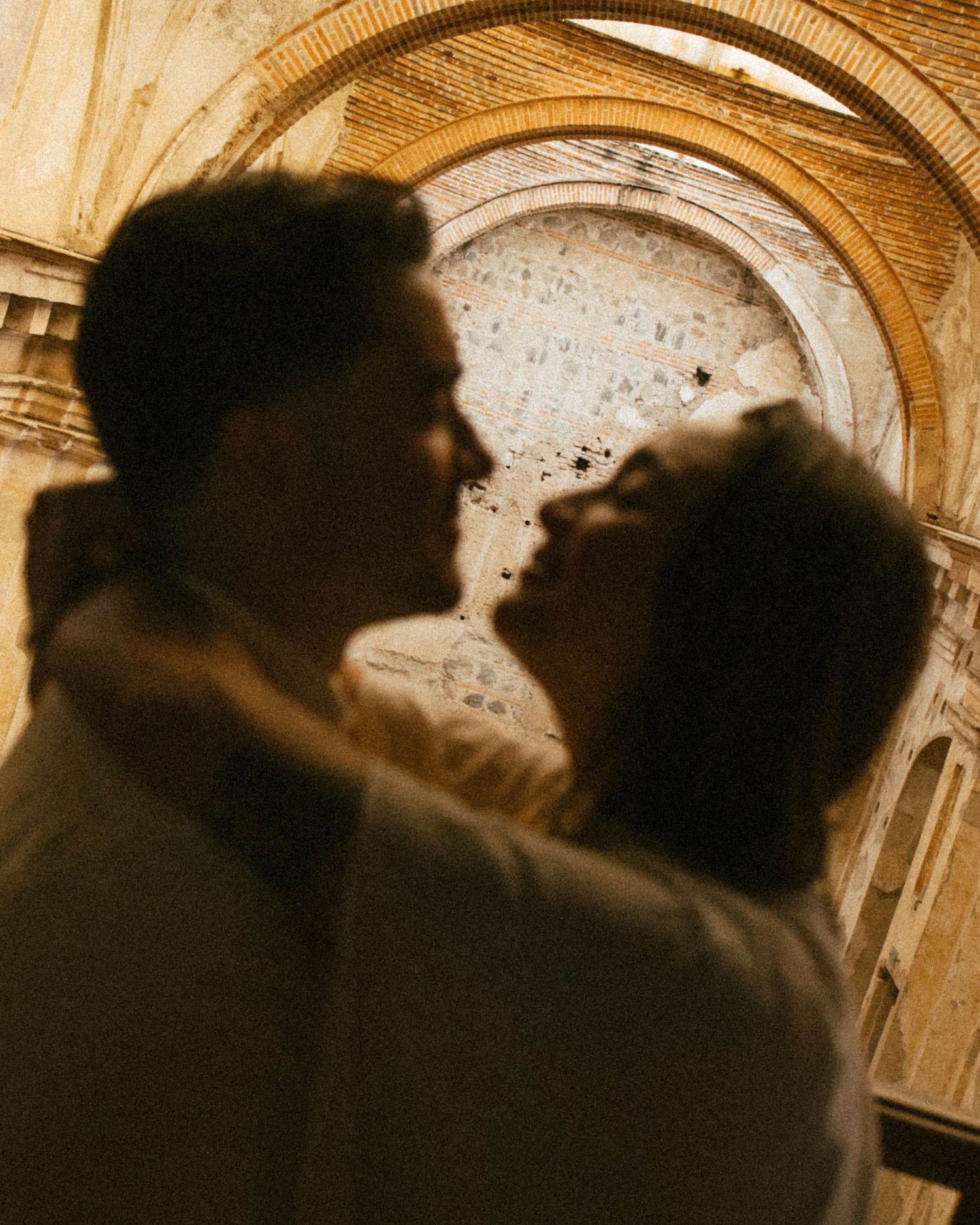 Romantic couple sharing an intimate moment kissing in front of ornate stone arched architecture.