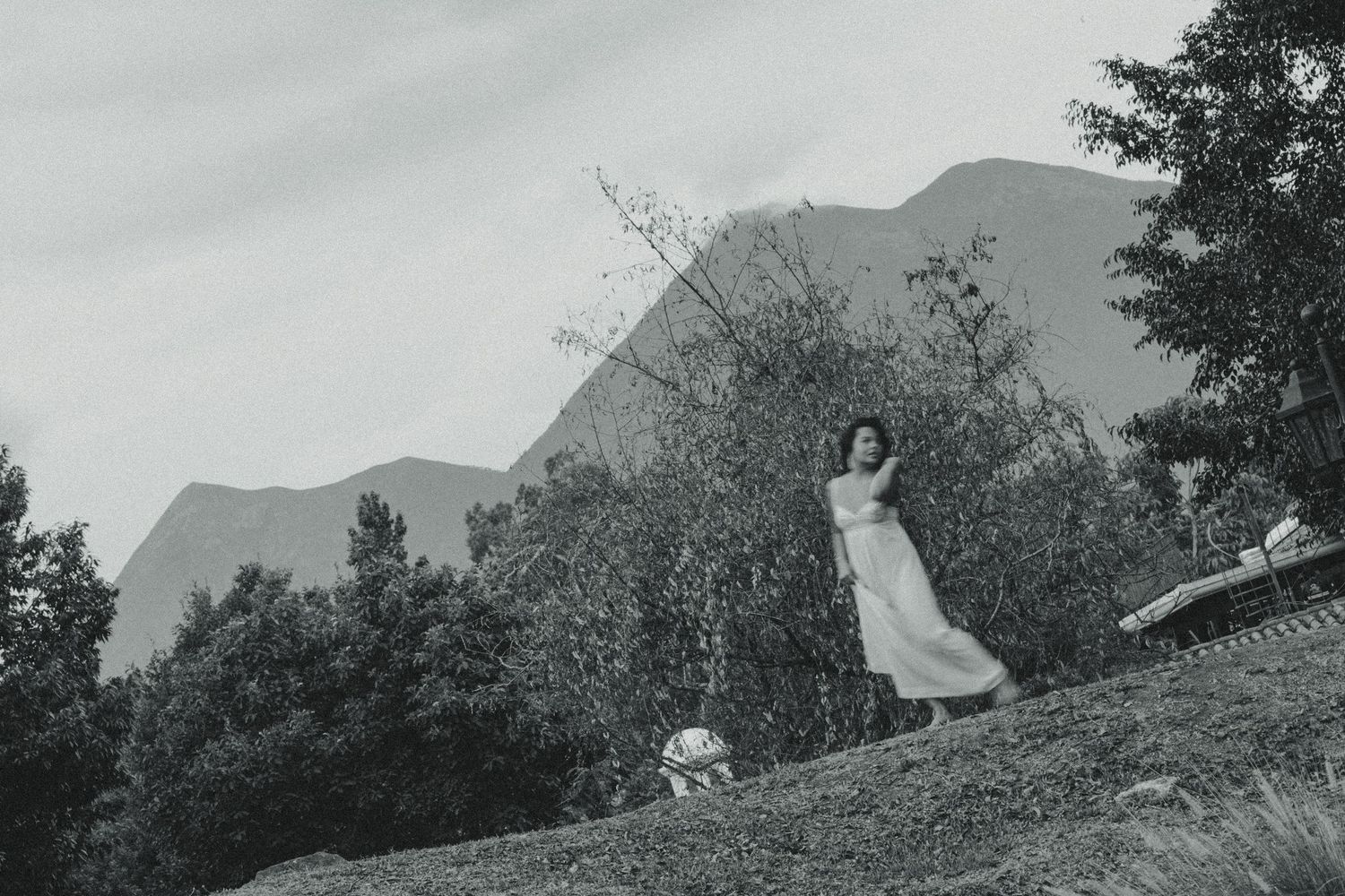 Black and white photo of a woman in a white dress standing on a hillside with mountains and trees behind her.