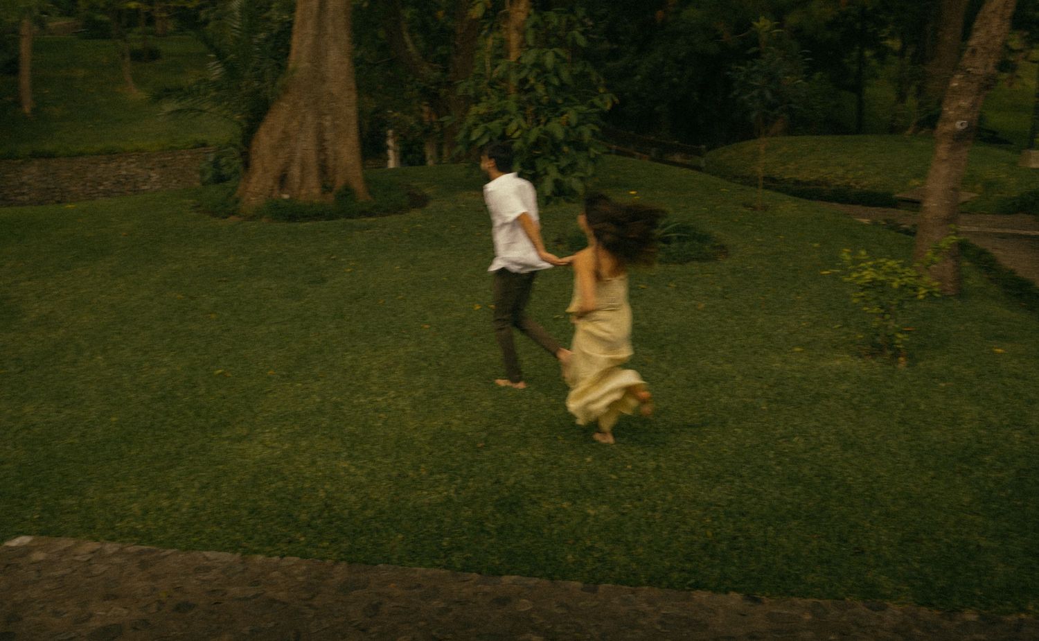 Couple holding hands running playfully on lush green lawn surrounded by tropical trees.