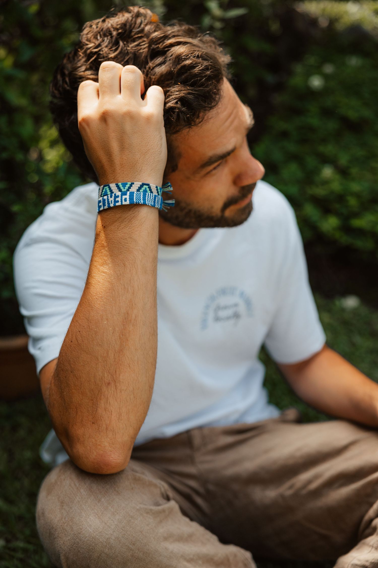 Bearded man in white t-shirt sitting outdoors wearing a blue woven bracelet, hand raised to head.