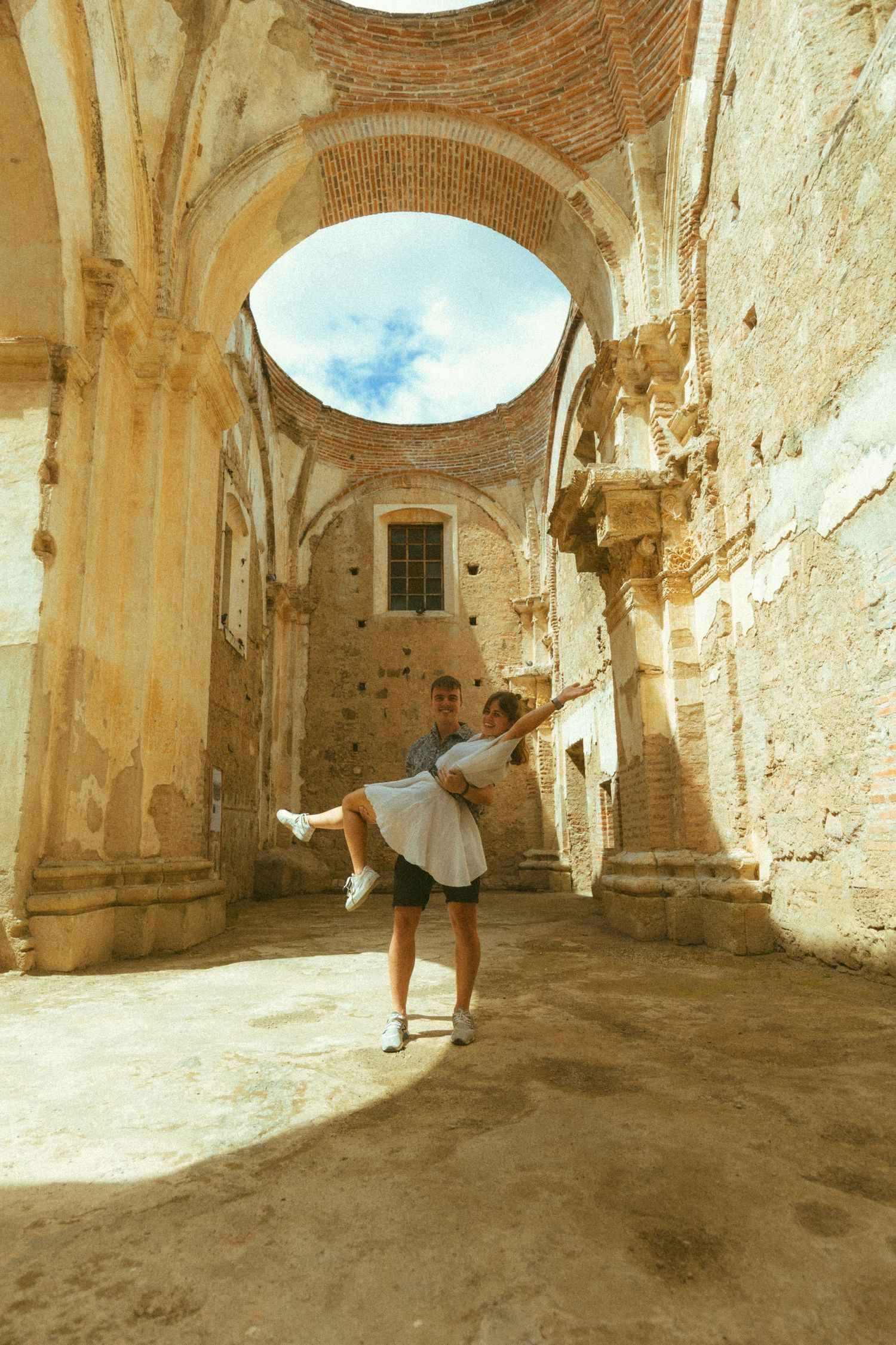 Couple dancing joyfully inside ancient stone ruins with arched ceiling open to blue sky.
