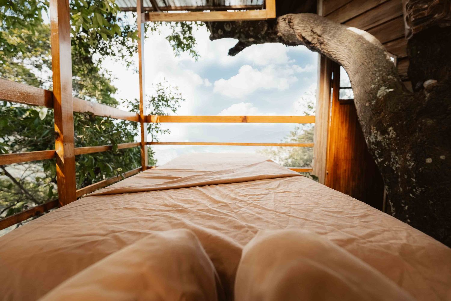 Cozy treehouse bed with wooden frame overlooking lush tropical trees and blue sky through open balcony.