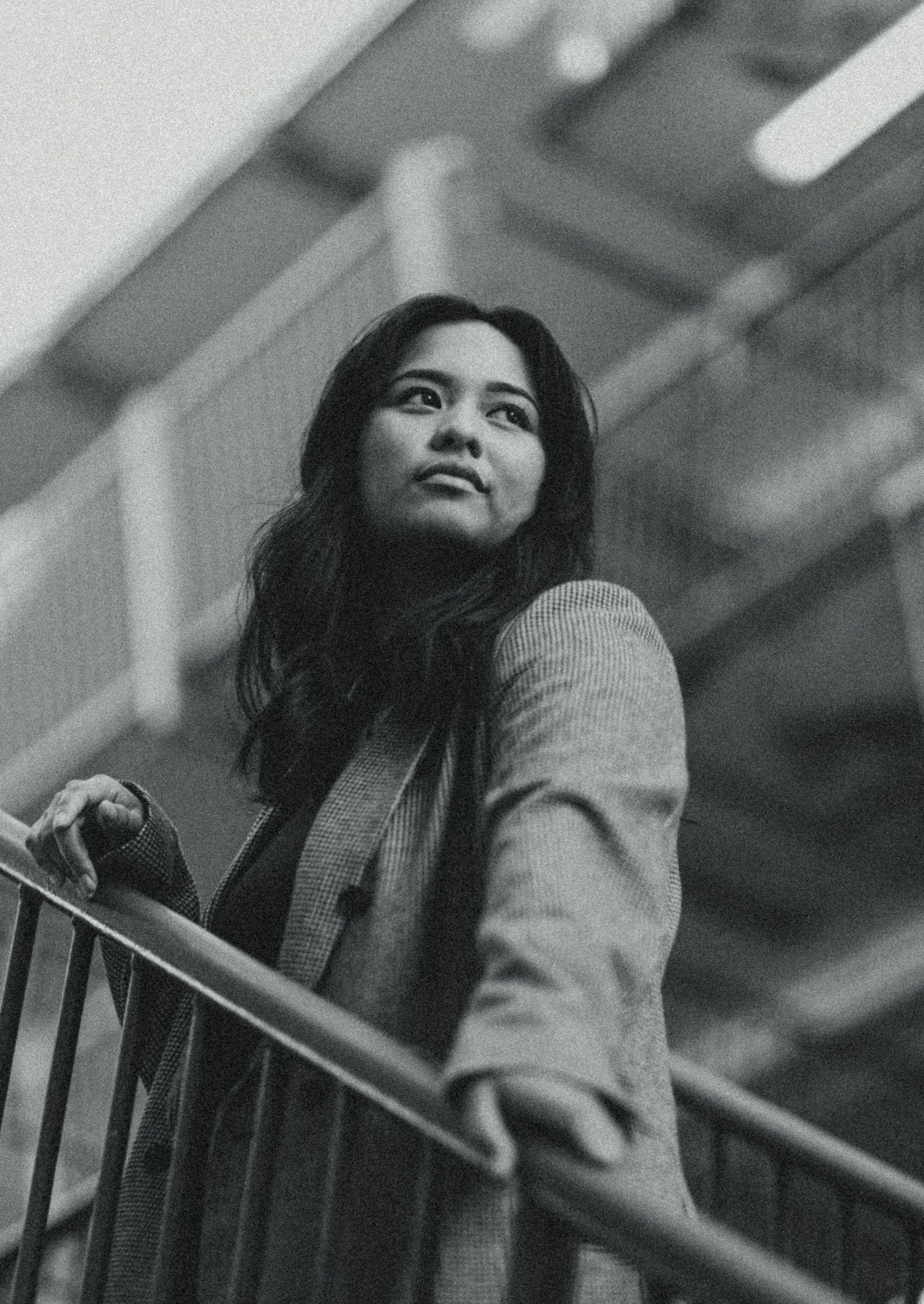 Black and white photo of a young woman in a blazer gazing upward while leaning on a railing outdoors.