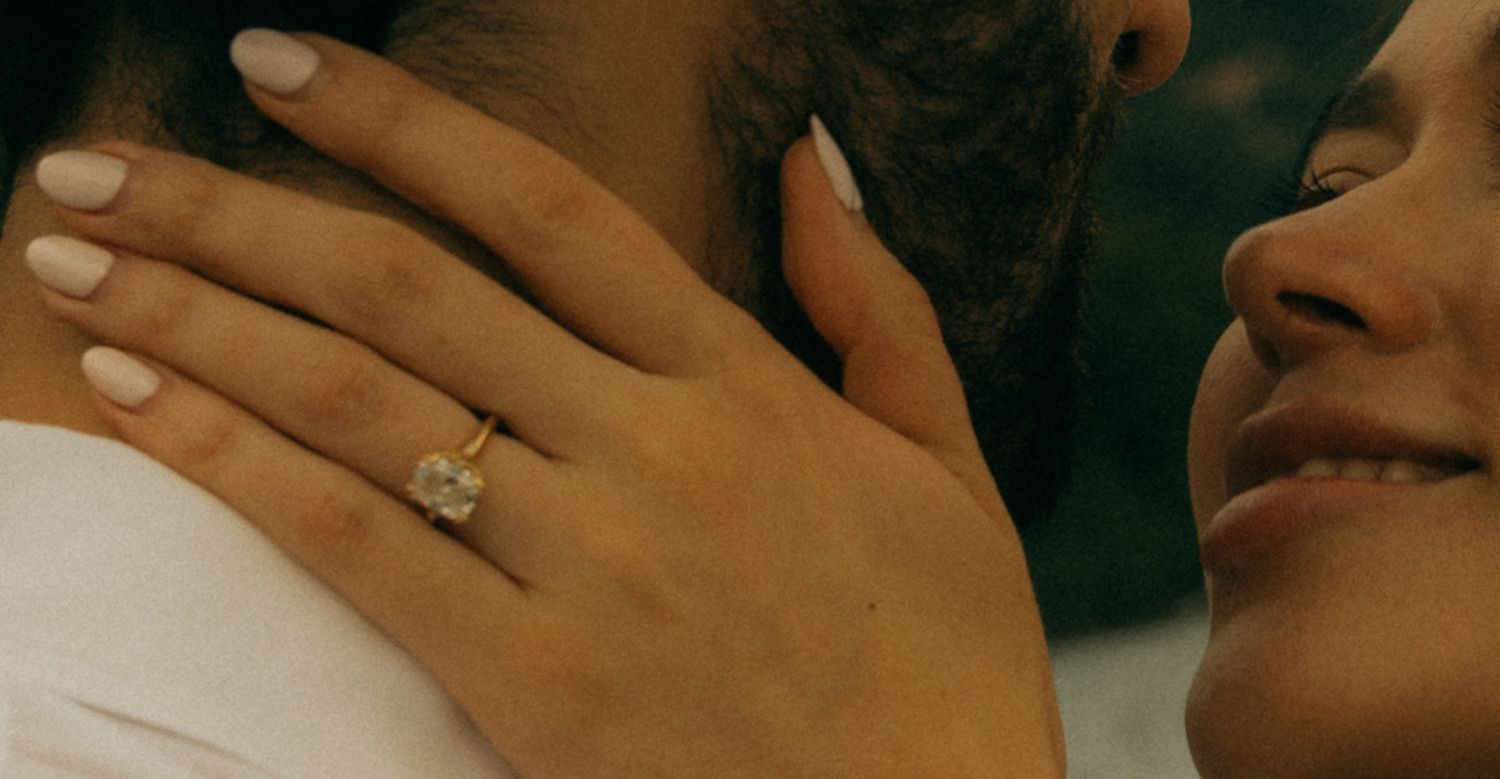 Close-up of a woman's hand with a diamond engagement ring resting on a man's shoulder.