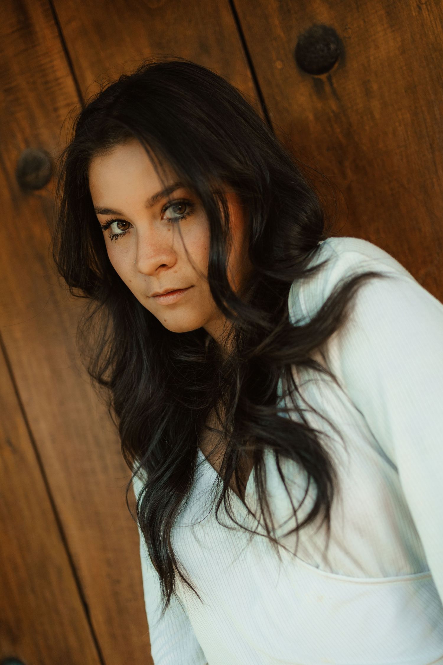 Young woman with long dark wavy hair wearing a white hoodie, posing against a rustic wooden door background.