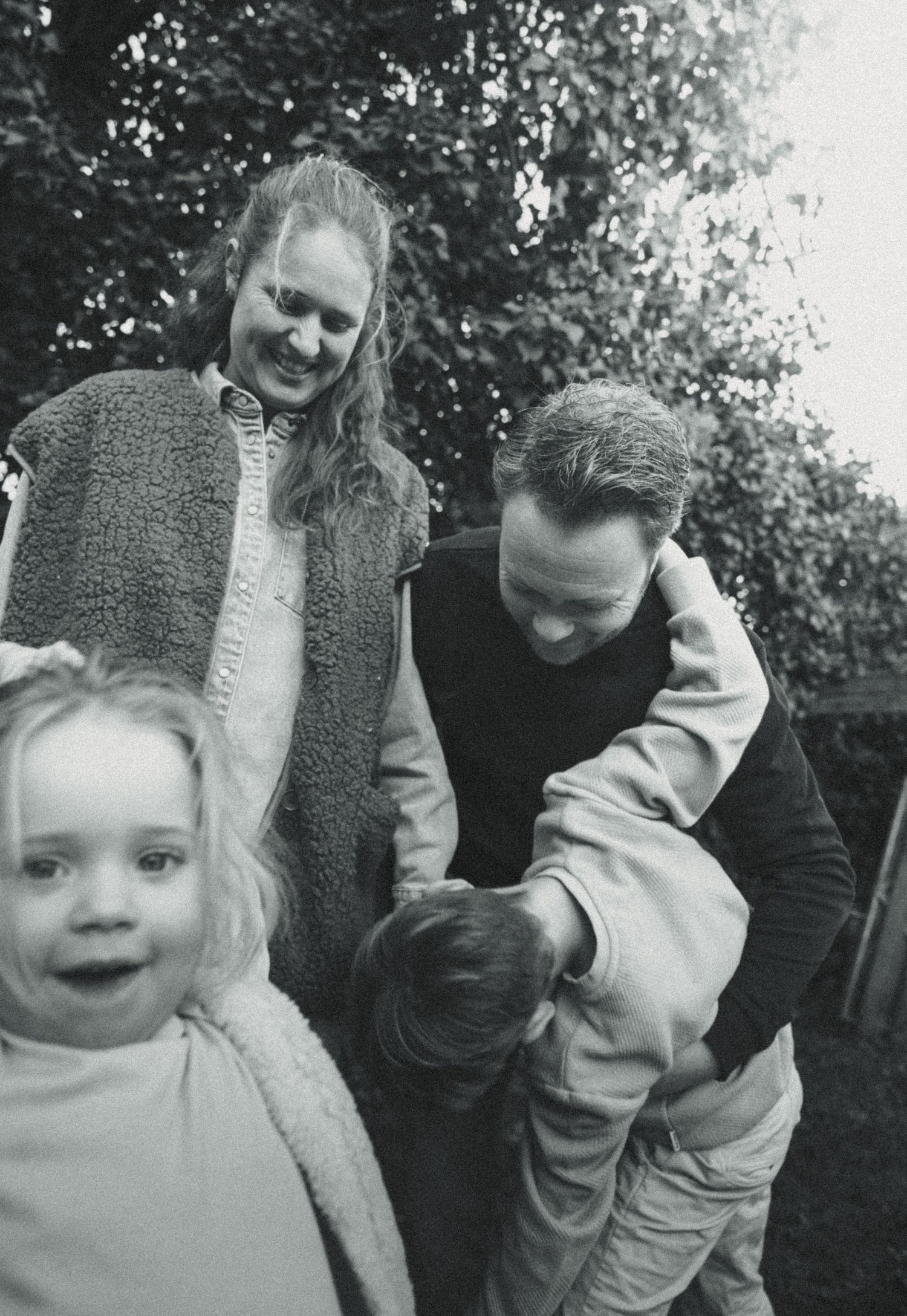 Black and white family photo with two adults smiling at two young children outdoors near trees.