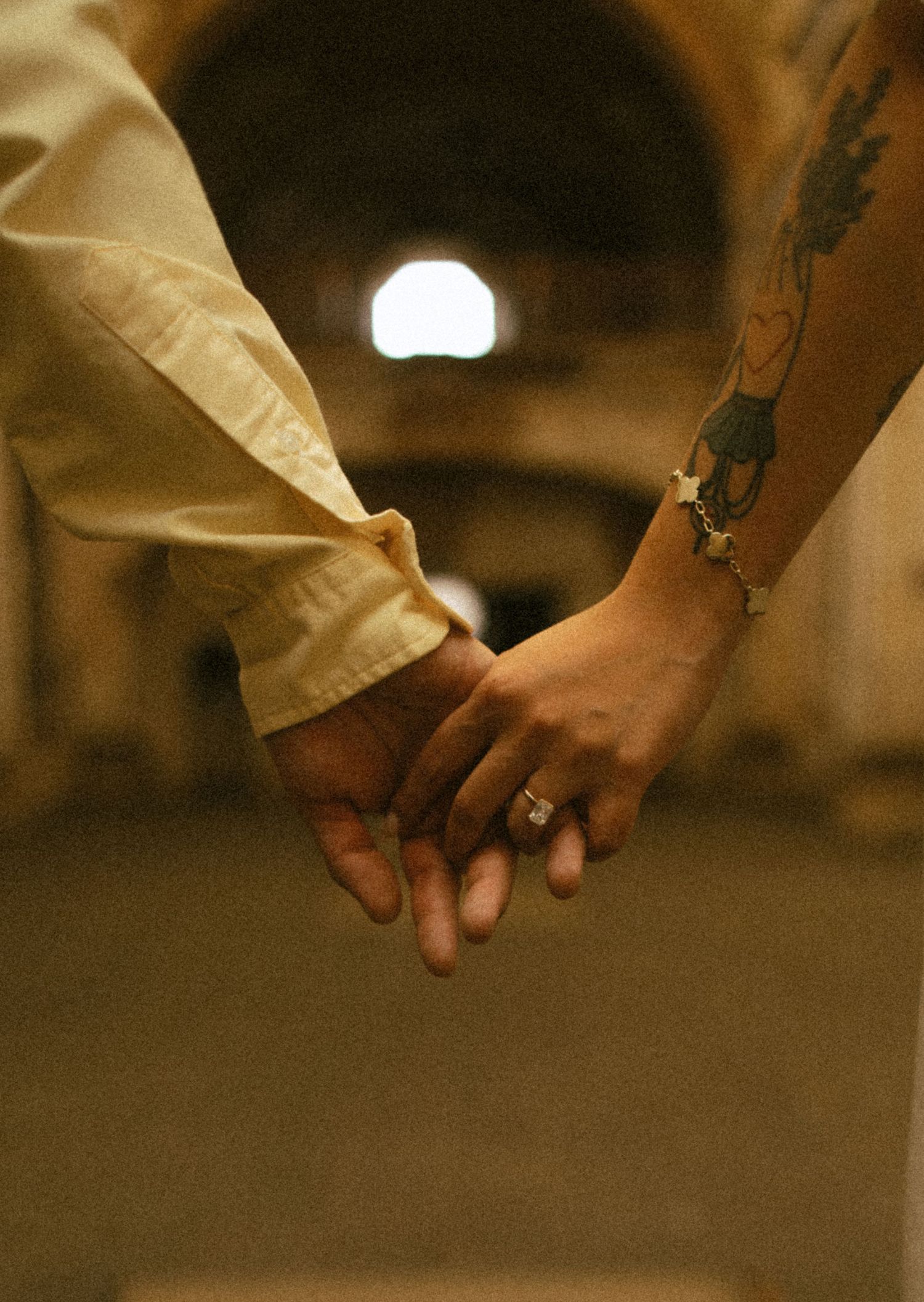 Couple holding hands with engagement ring and floral tattoo visible, standing in a dimly lit archway.