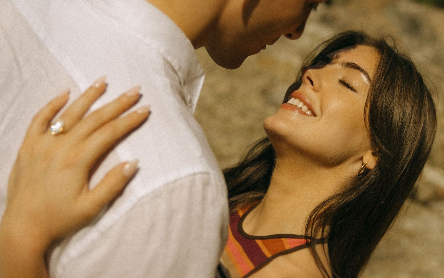 A smiling young woman gazes up at a man while embracing outdoors, wearing a ring on her finger.