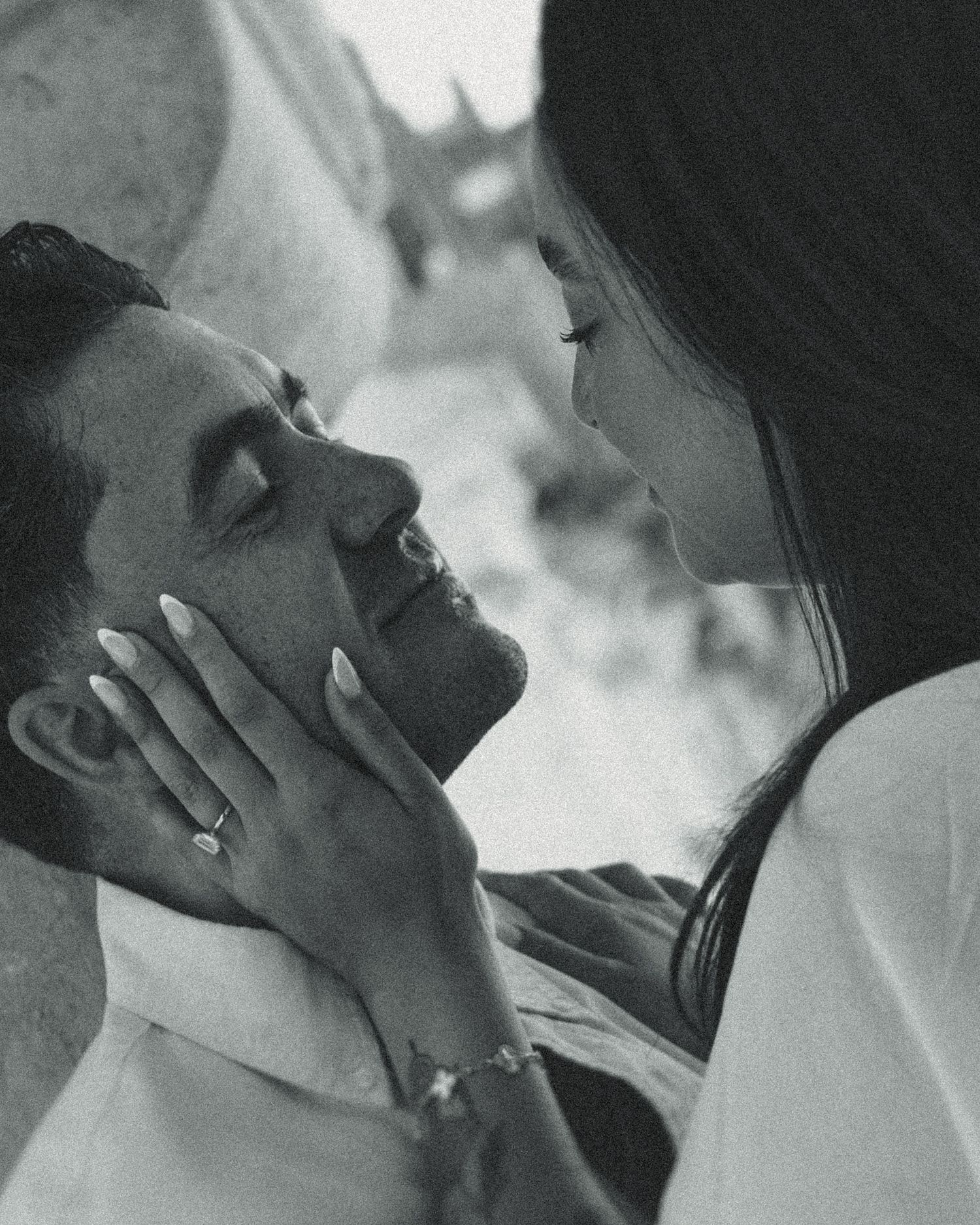 Black and white photo of a woman gently cradling a smiling man's face, her ringed hand on his cheek, in a tender romantic moment.