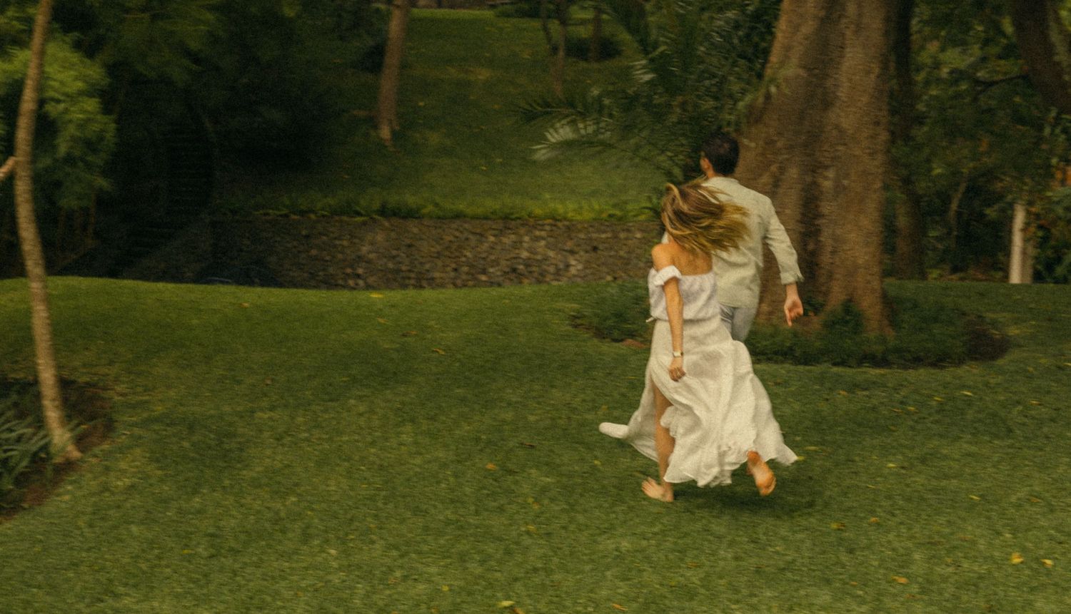 Two people in white clothing run across a lush green lawn surrounded by tall trees in a dreamy outdoor scene.