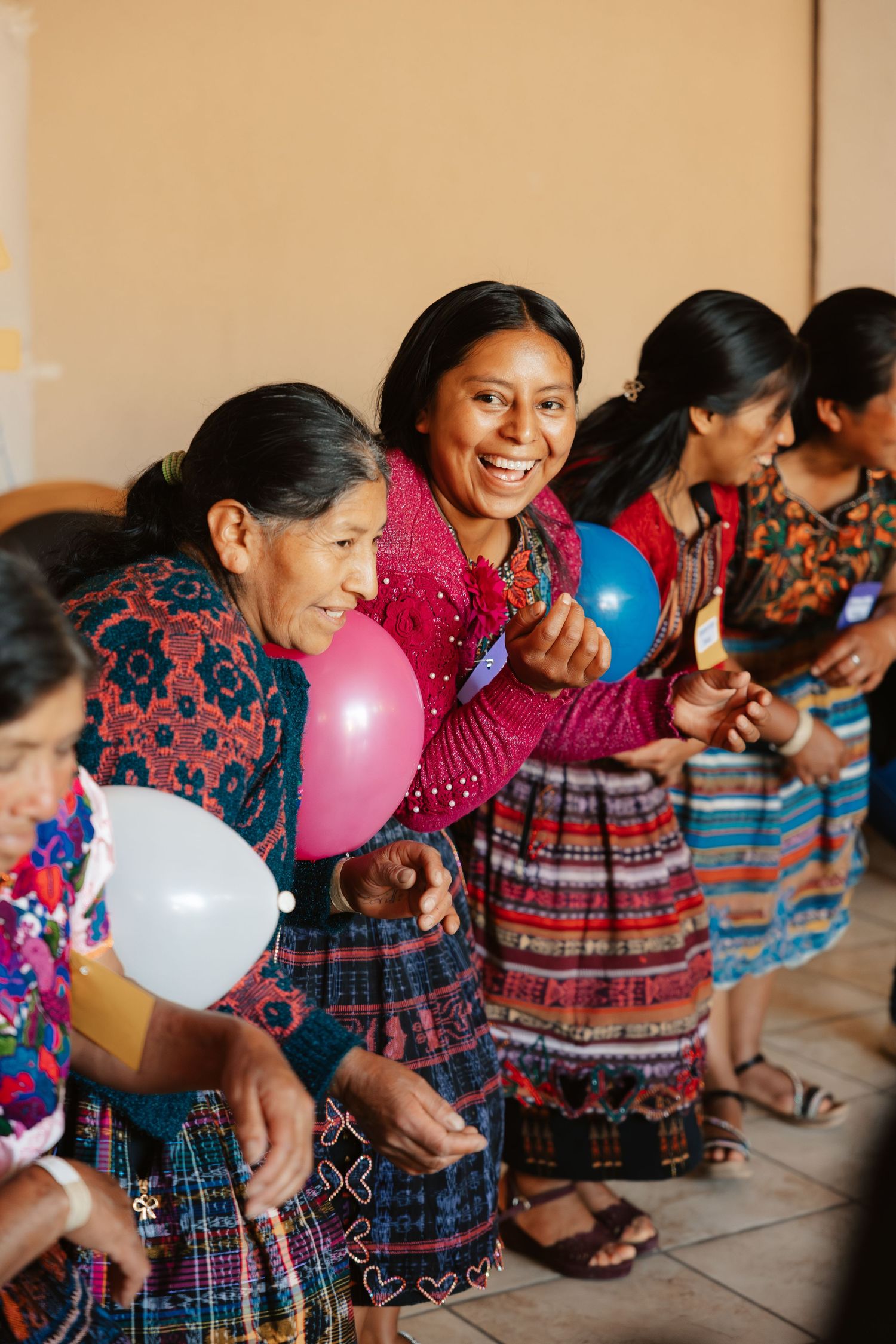 Indigenous Guatemalan women in colorful traditional huipil dresses laugh and hold balloons during a festive cultural celebration.
