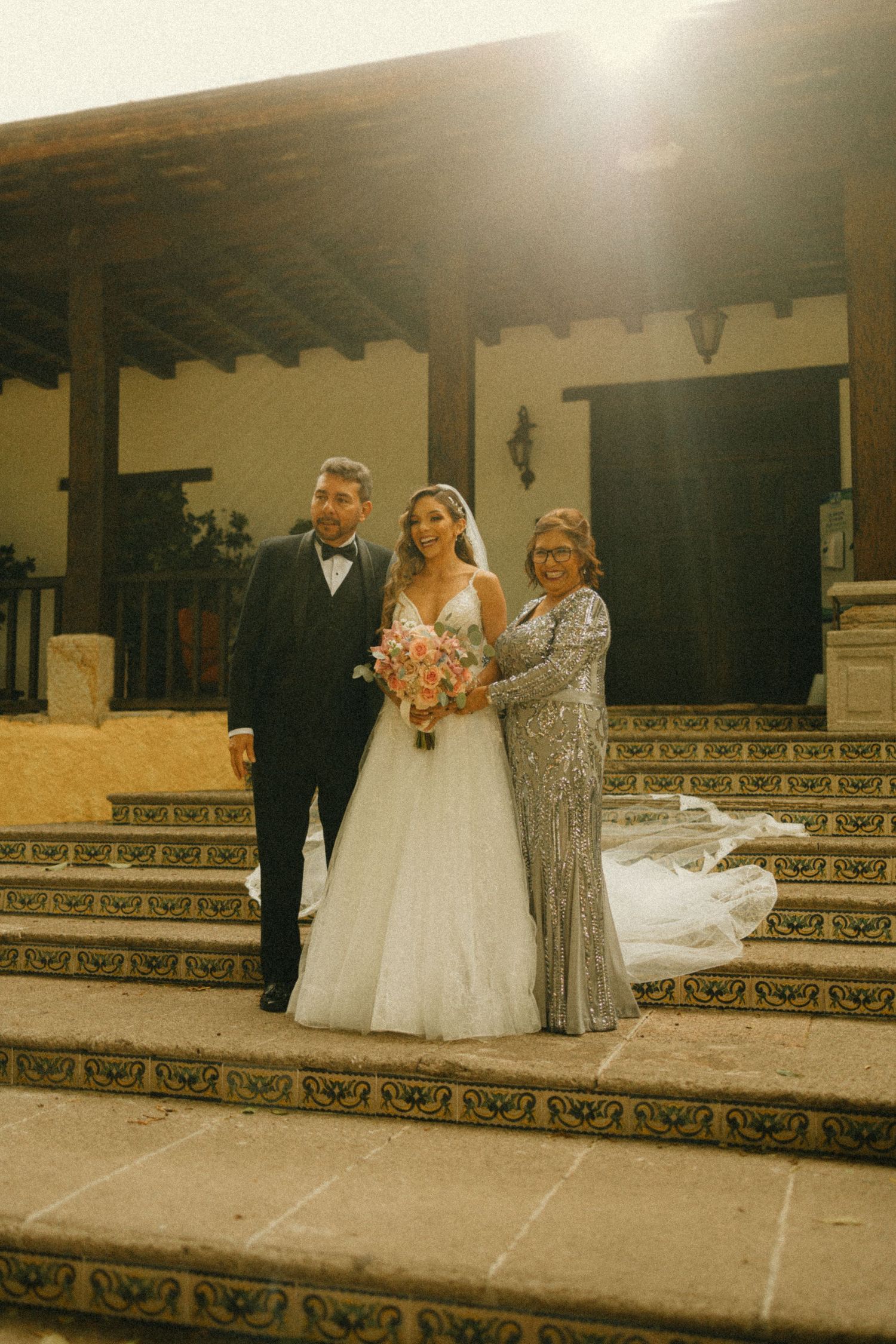 Bride in white gown with groom and mother on tiled steps at golden hour wedding venue.