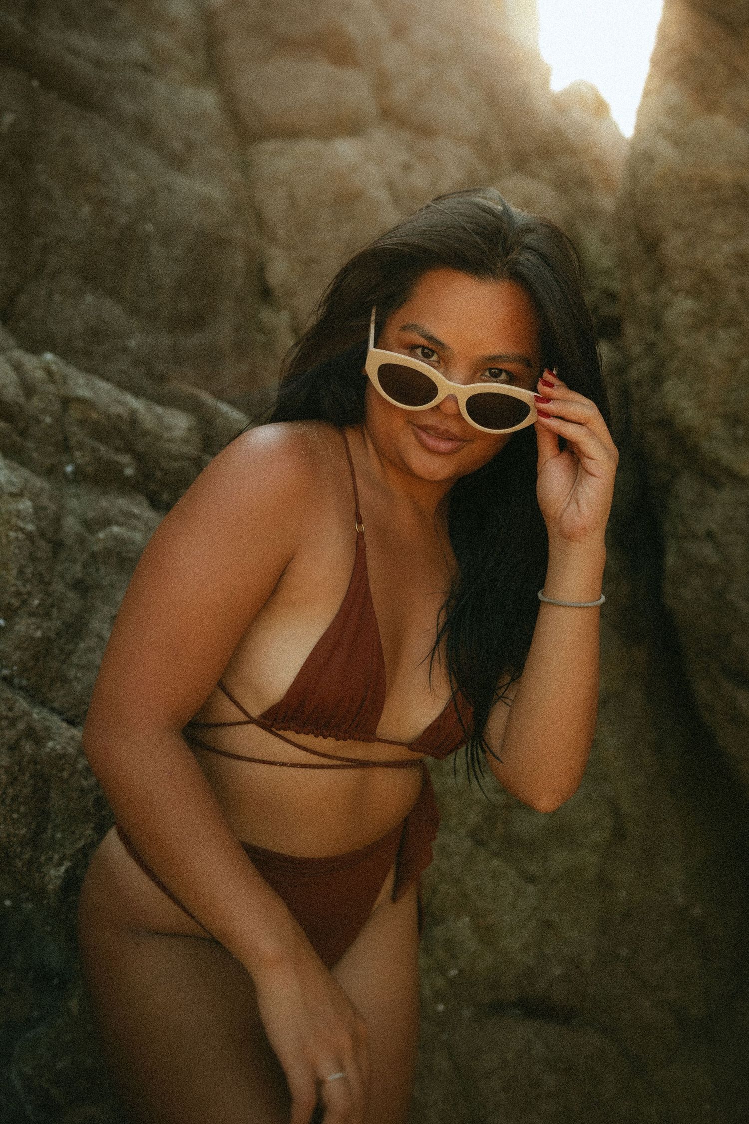 Woman in brown bikini posing between rocks, lowering white cat-eye sunglasses, with warm golden backlight.