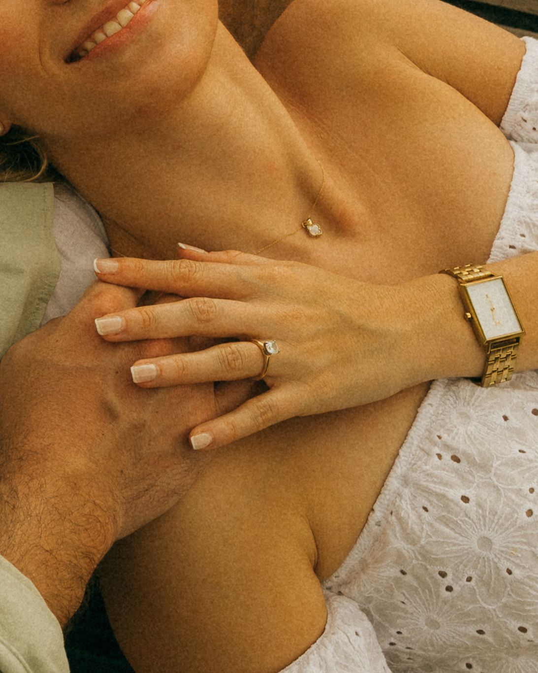 Close-up of a woman's hand wearing a diamond ring and gold watch, resting on a white eyelet dress.