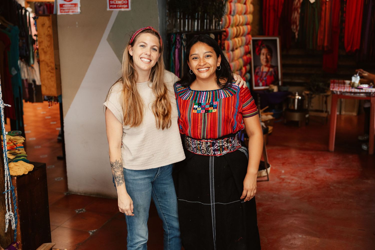 Two women smiling together in a colorful textile shop, one wearing traditional Guatemalan huipil embroidered blouse.