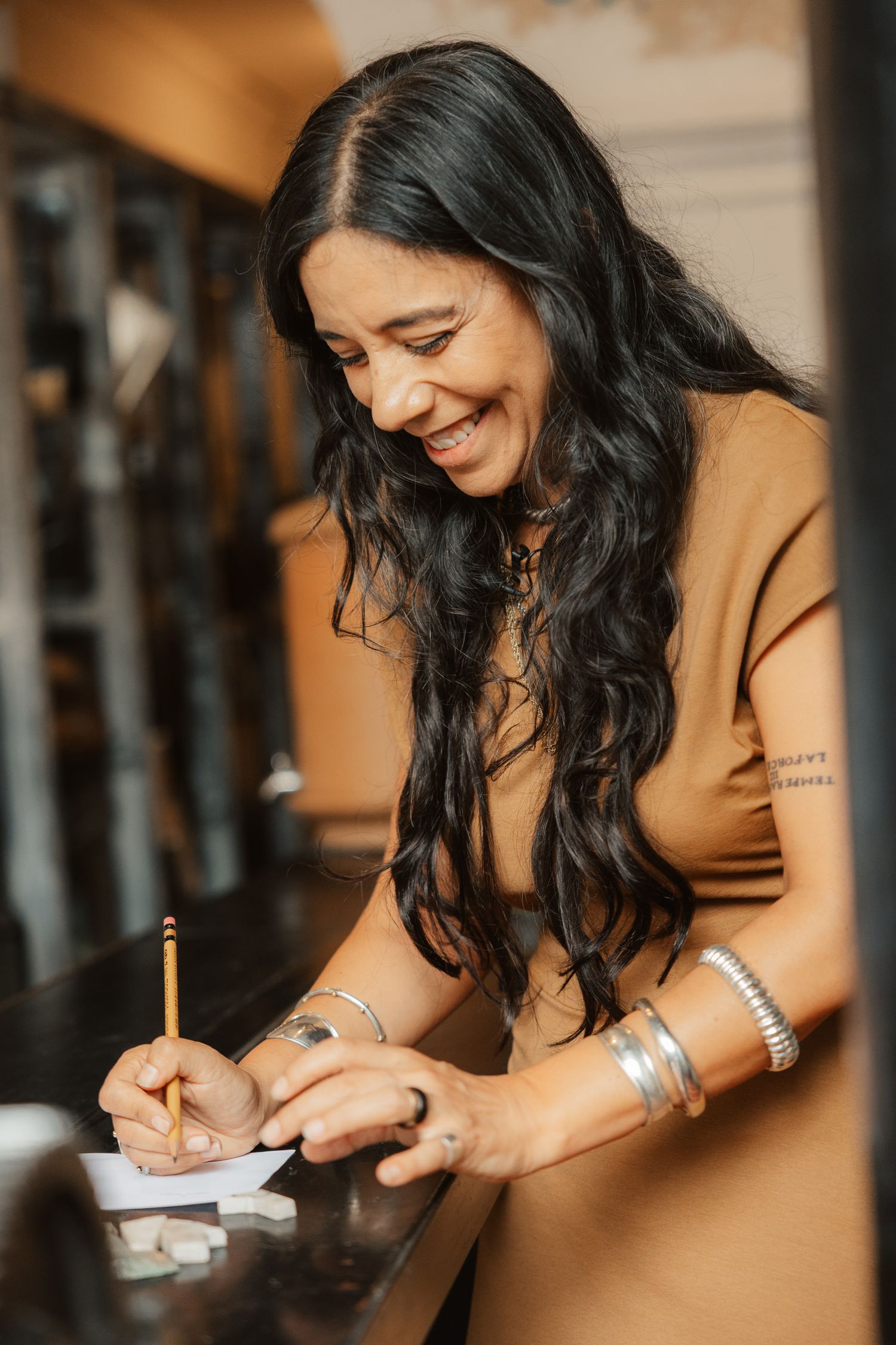 Smiling woman with long dark hair writing with a pencil, wearing a tan dress and silver bracelets.